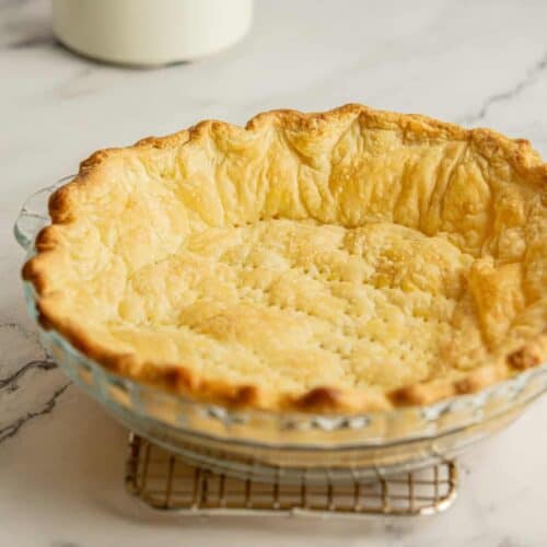 A golden brown pie crust in a glass dish sits on a cooling rack on a marble countertop. The crust is flaky and evenly baked, with a gently crimped edge.