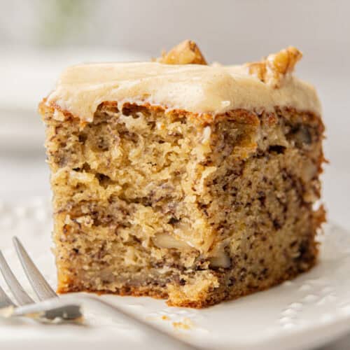 A close-up of a single slice of banana walnut cake with frosting on top, served on a white plate. The cake appears moist and topped with small nut pieces, with a fork resting beside it. The background is soft and blurred.