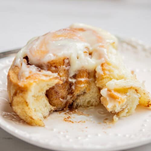 Close-up of a partially eaten cinnamon roll on a white plate. It is covered with creamy icing, highlighting the fluffy texture and warm, cinnamon fillings inside.