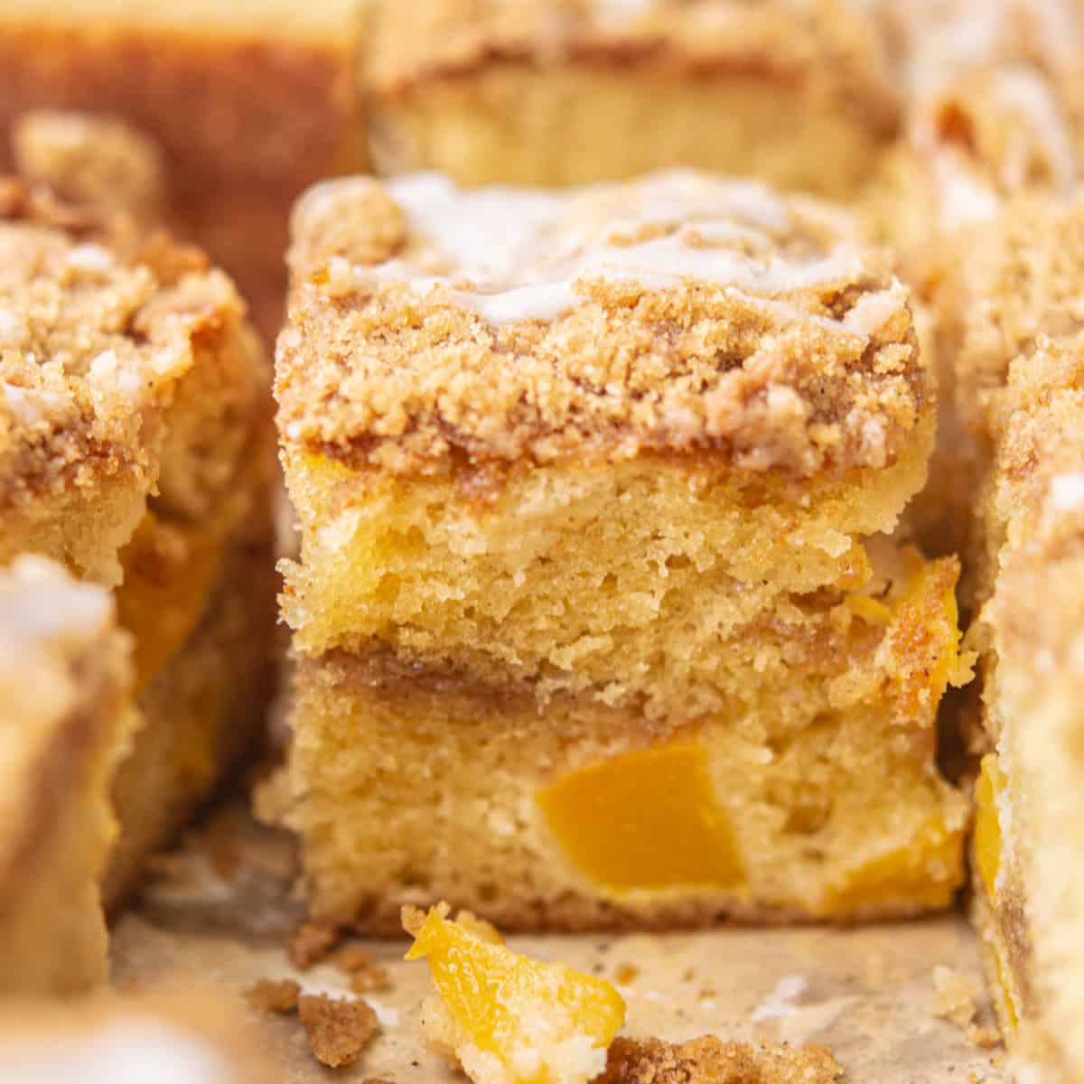 Close-up of a slice of crumbly peach coffee cake topped with streusel and drizzled with icing. The cake has visible chunks of peach and a golden-brown crust, creating a warm and inviting appearance.