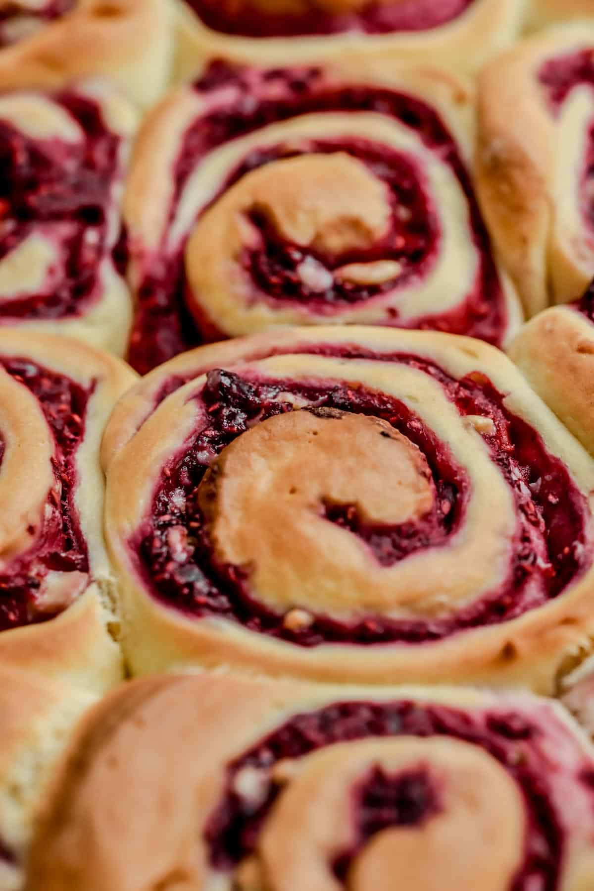 Close-up of freshly baked Raspberry Rolls, showing golden-brown swirled dough filled with vibrant red raspberry filling. The Raspberry Rolls are closely packed together, highlighting their soft, fluffy texture.
