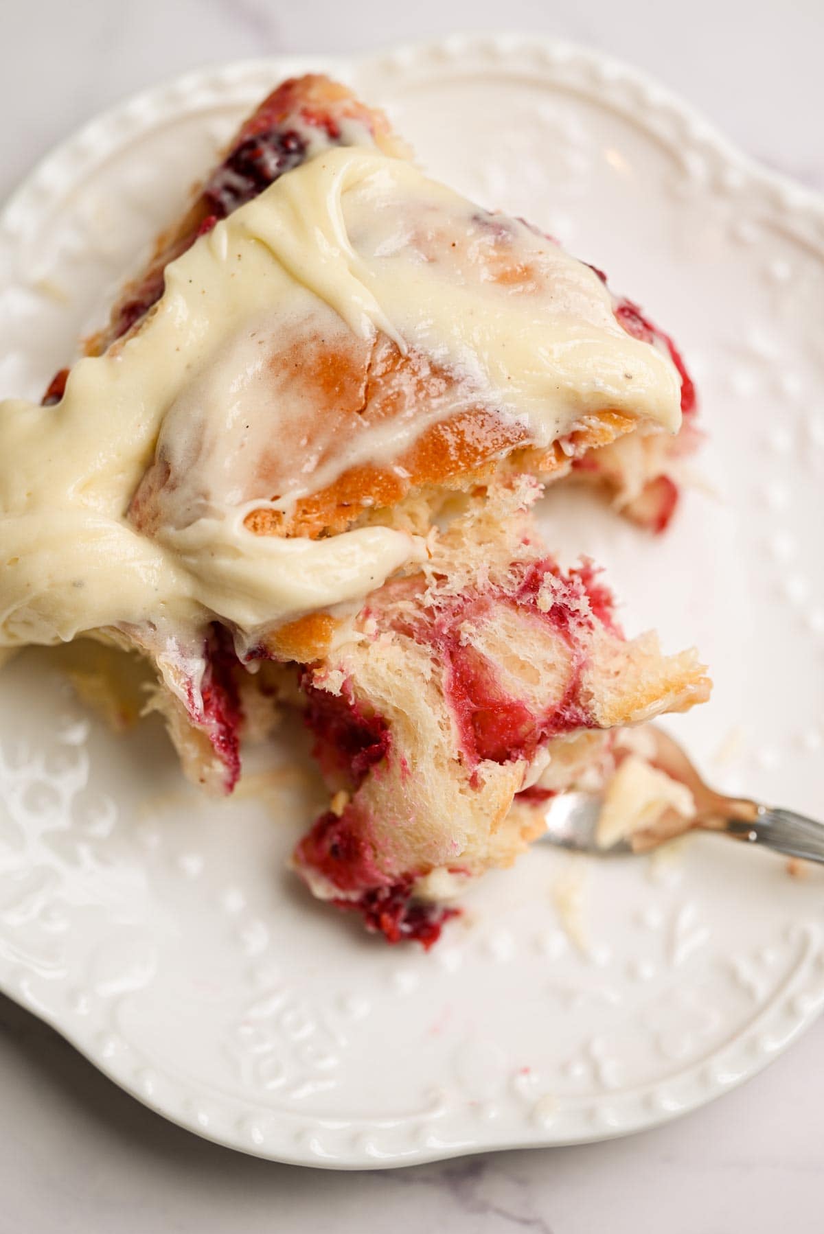 A close-up of Raspberry Rolls with sweet raspberry filling and creamy frosting on a decorative white plate, as a fork takes a bite from the delicious pastry.