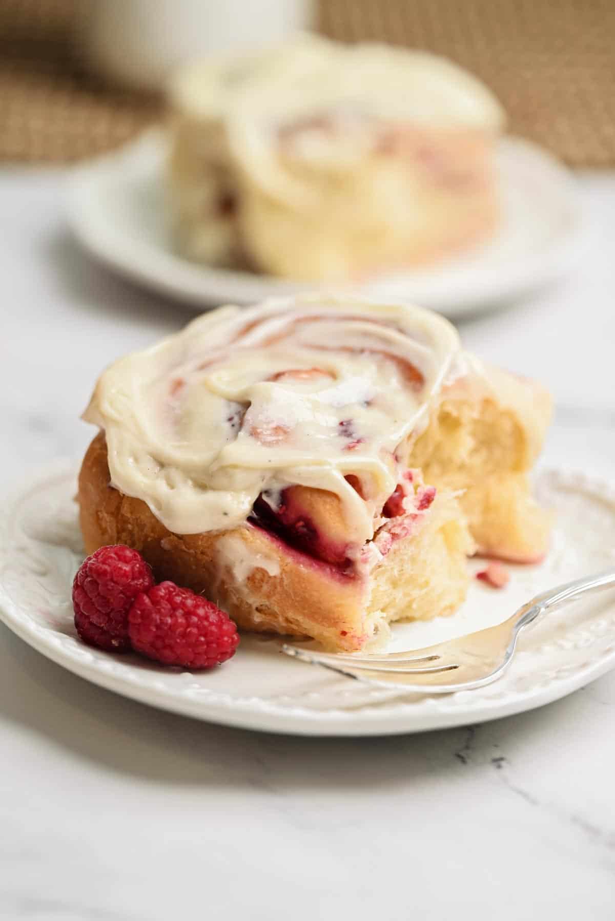 A Raspberry Rolls sweet roll topped with cream cheese frosting sits on a white plate, garnished with fresh raspberries. A fork rests beside the roll, while another Raspberry Roll is blurred in the background.