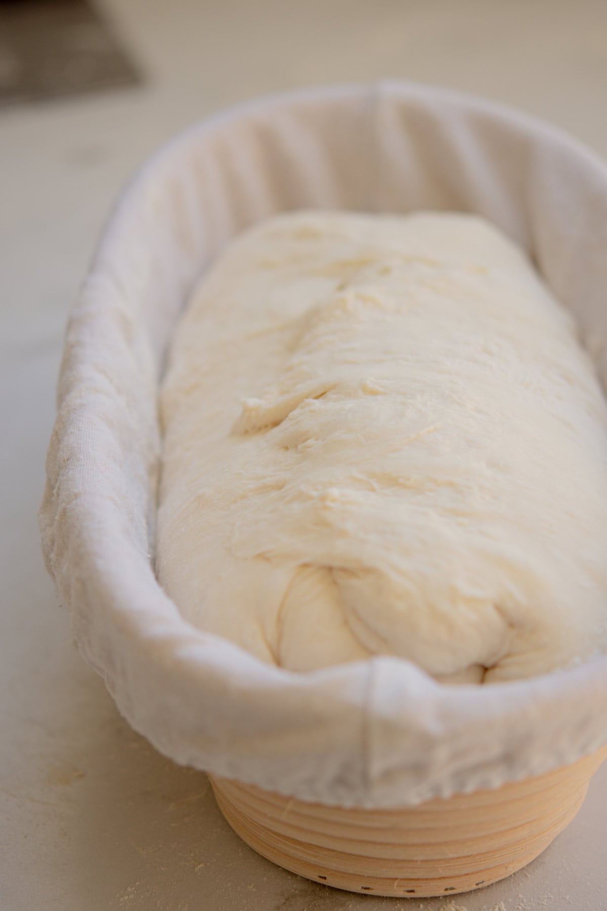 A loaf of risen dough rests in an oval-shaped, cloth-lined proofing basket on a white surface, ready for baking.