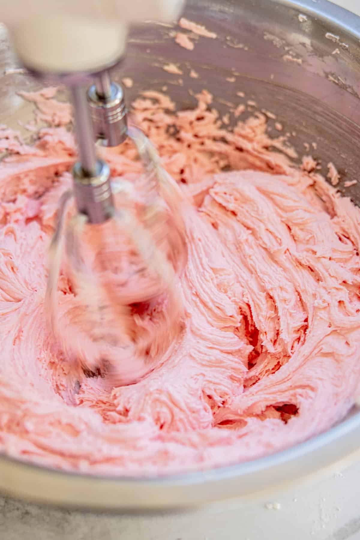 A close-up of a mixing bowl filled with pink frosting being blended by an electric mixer. The frosting appears smooth and creamy as the beaters swirl through it. The bowl is metallic and resting on a white countertop.