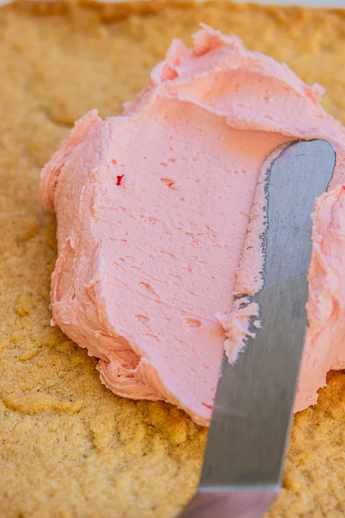 A knife spreading pink frosting over a baked, golden-brown surface.