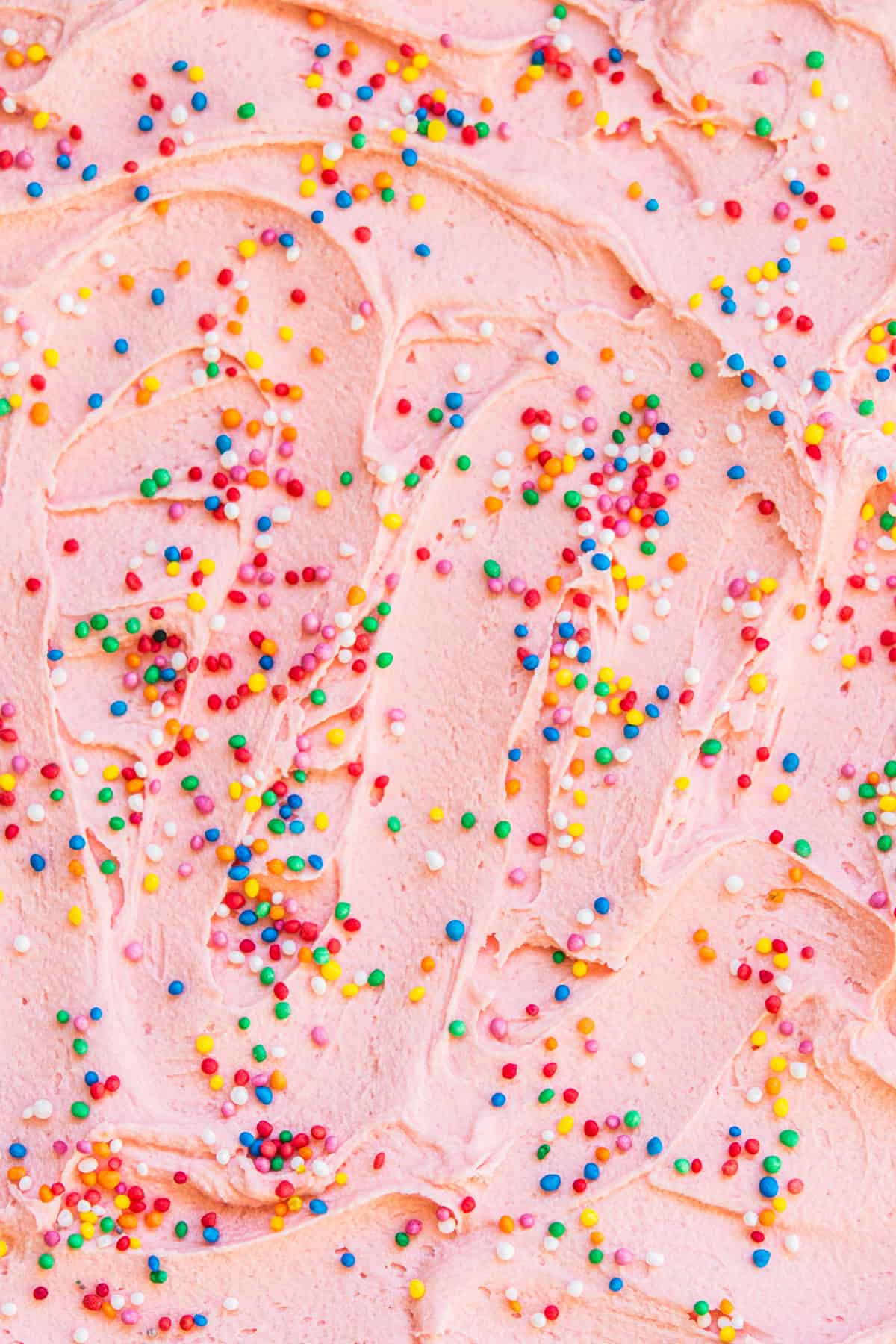 Close-up of pink frosting with colorful sprinkles scattered on top. The texture is smooth with swirled patterns, and the sprinkles add a playful, vibrant touch.