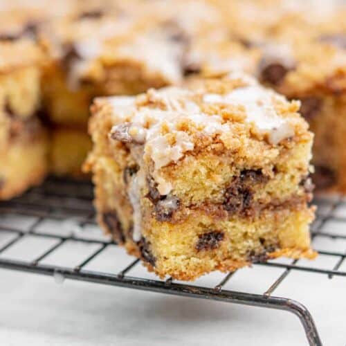 A close-up of a square piece of chocolate chip coffee cake with a crumbly topping and chocolate chips, resting on a cooling rack. The cake appears moist with layered textures and a light glaze drizzle on top. Other pieces are blurred in the background.