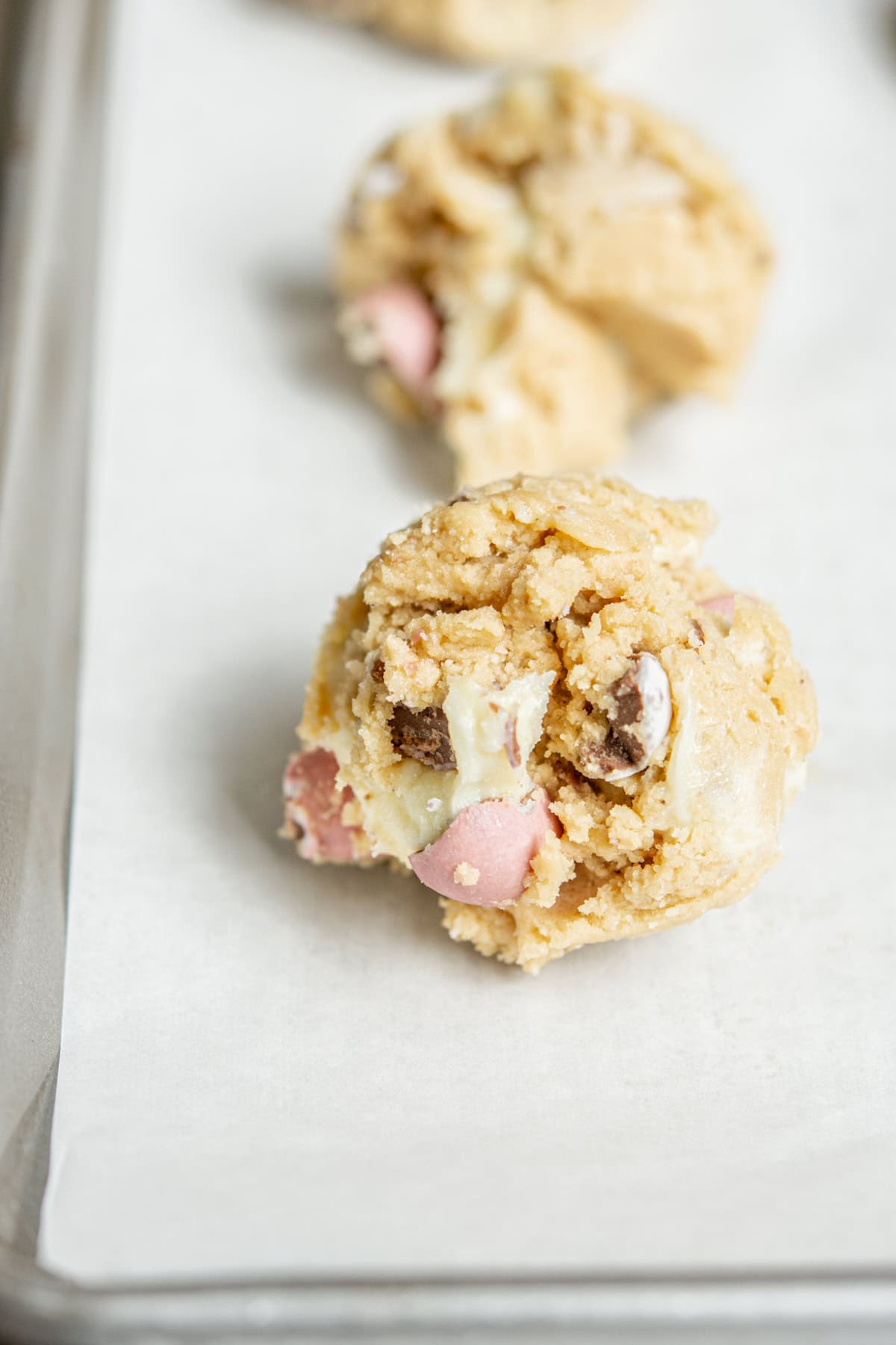 Close-up of cookie dough balls with chunks of chocolate spread on a parchment-lined baking sheet. The dough appears ready to bake, featuring pieces of pink and white chocolate.