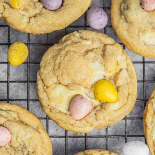 Easter cheesecake cookies topped with pastel-colored candy eggs are cooling on a wire rack. Their light brown color and soft texture are beautifully showcased, offering a delightful sugary surface and festive charm.