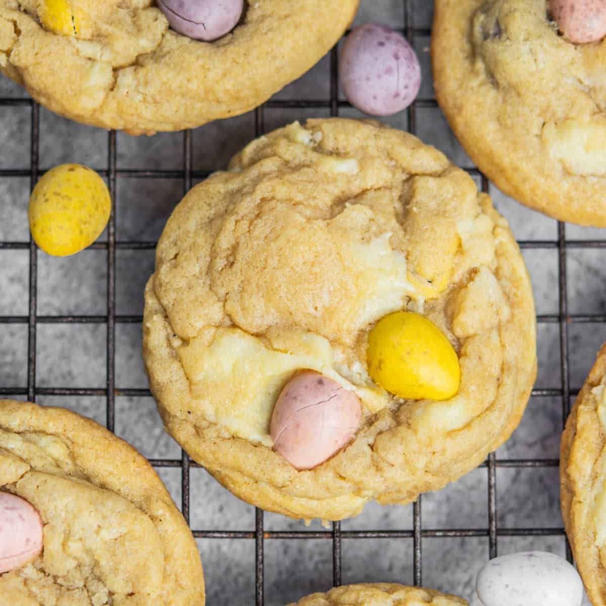 Easter cheesecake cookies topped with pastel-colored candy eggs are cooling on a wire rack. Their light brown color and soft texture are beautifully showcased, offering a delightful sugary surface and festive charm.