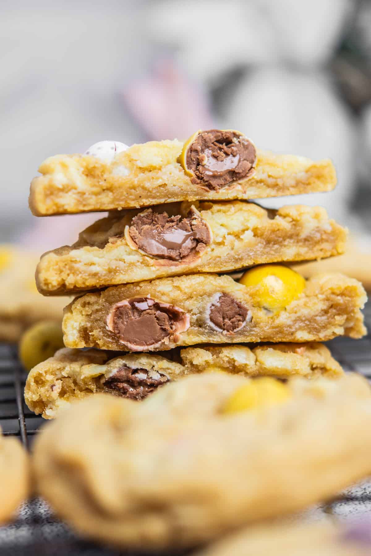 A stack of Easter cookies, cut in half to reveal colorful chocolate candies inside. They boast a golden-brown exterior and soft interior, echoing a cheesecakes creamy allure. Placed on a wire rack, more treats are blurred in the background, tempting taste buds in anticipation.