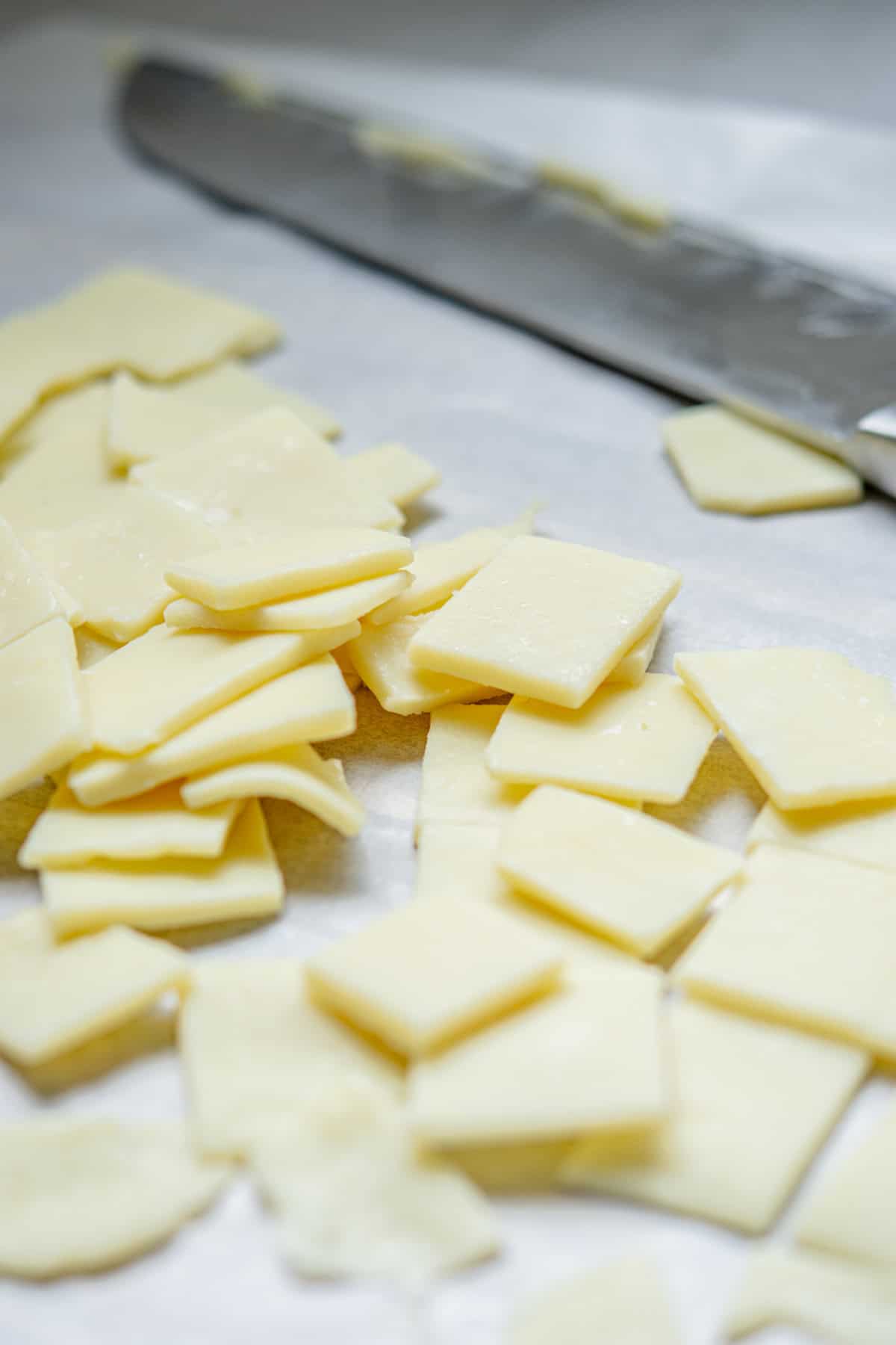 Slices of cheese on a sheet of parchment paper with a knife in the background. The cheese is cut into thin squares and irregular shapes.