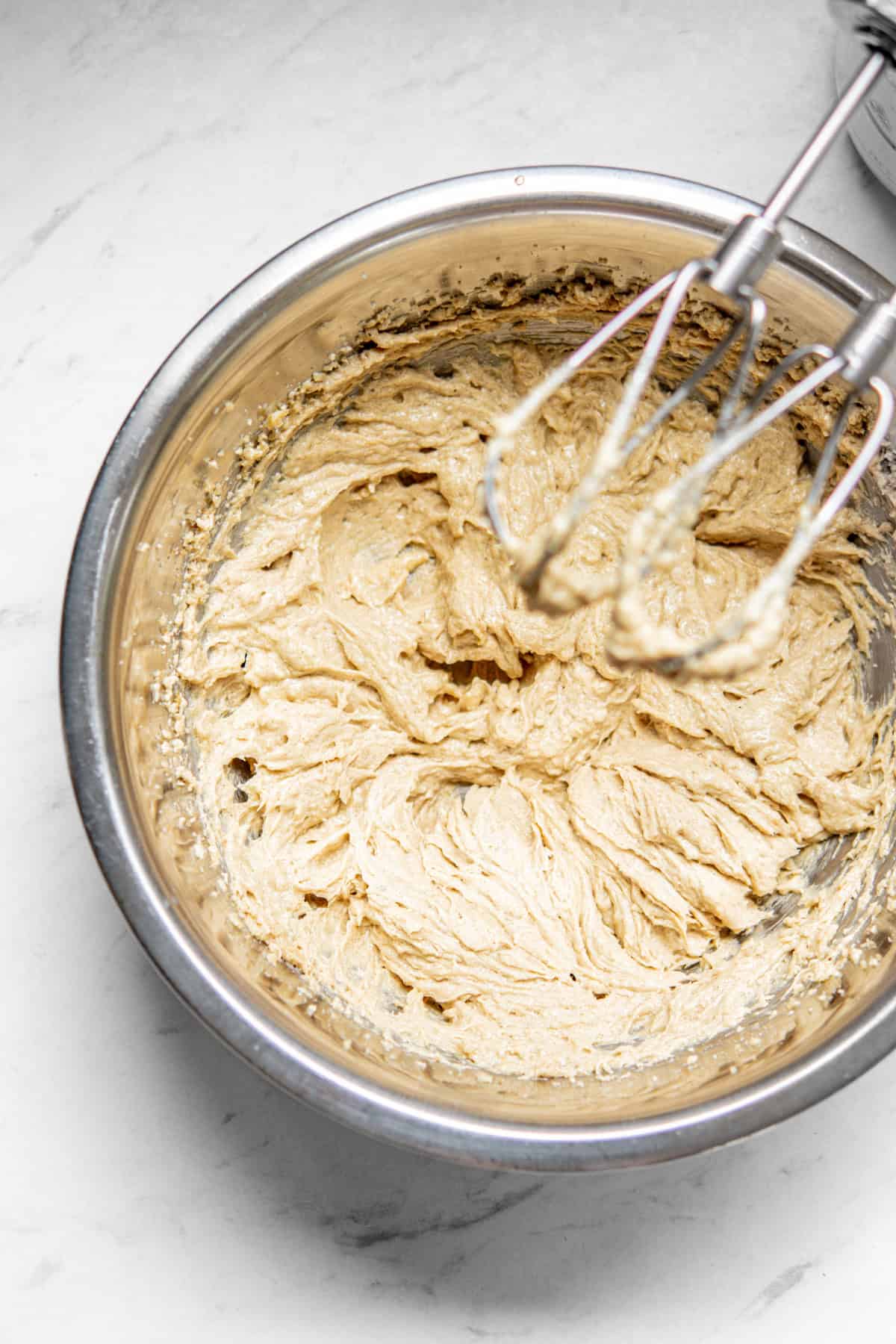 A stainless steel bowl containing a creamy, beige mixture is placed on a marble countertop. An electric hand mixer with two beaters rests above the bowl, indicating the mixture has been freshly prepared.
