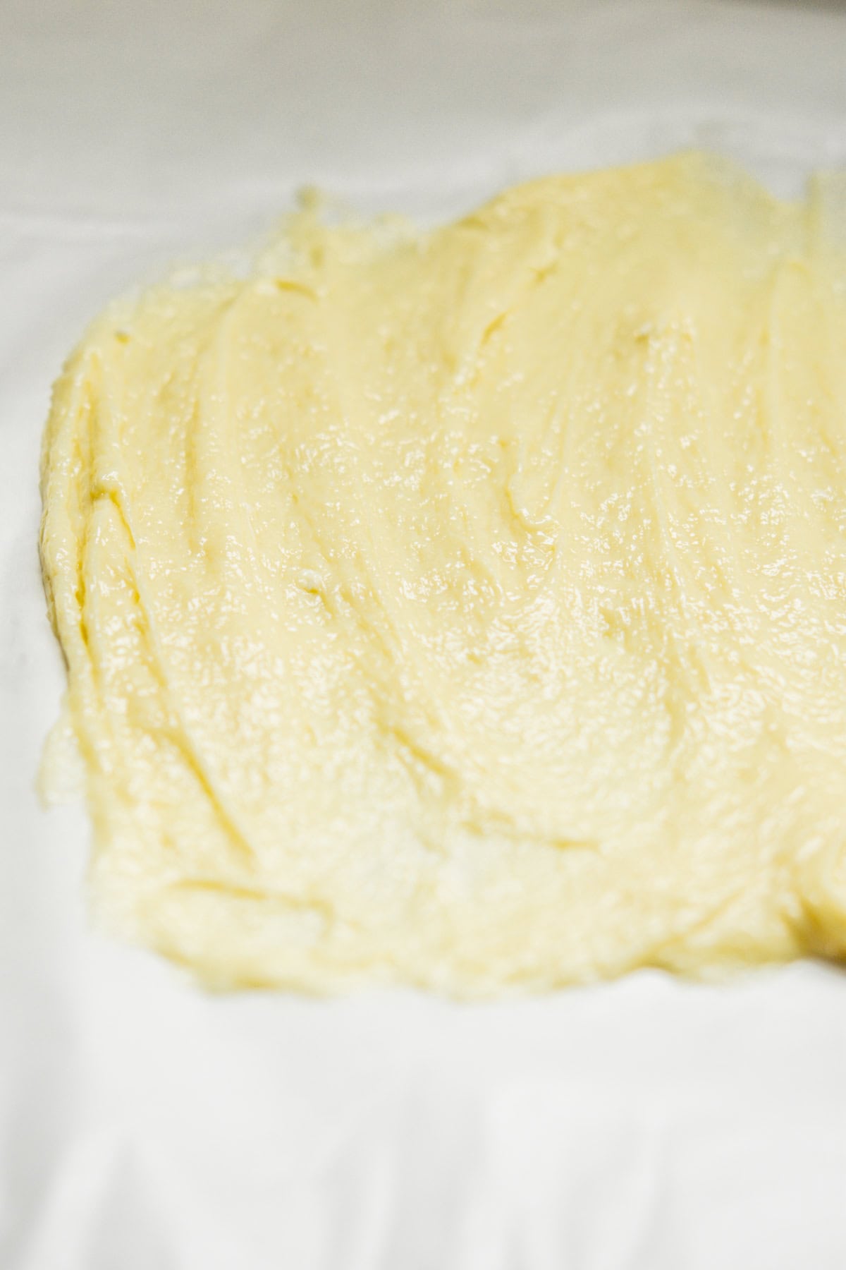A thick, creamy, pale yellow paste is spread unevenly on a white surface. The texture appears smooth and buttery, with visible swirls and ridges. The background is slightly blurred, emphasizing the texture of the paste.