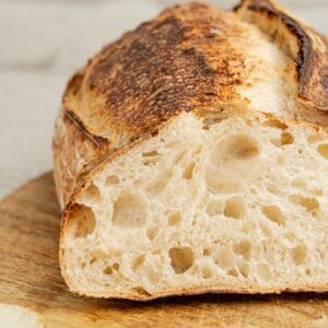 Close-up of a rustic artisan bread loaf with a golden-brown crust, sliced to show its airy, soft interior. The bread rests on a wooden surface, highlighting the texture and color contrast between the crust and crumb.