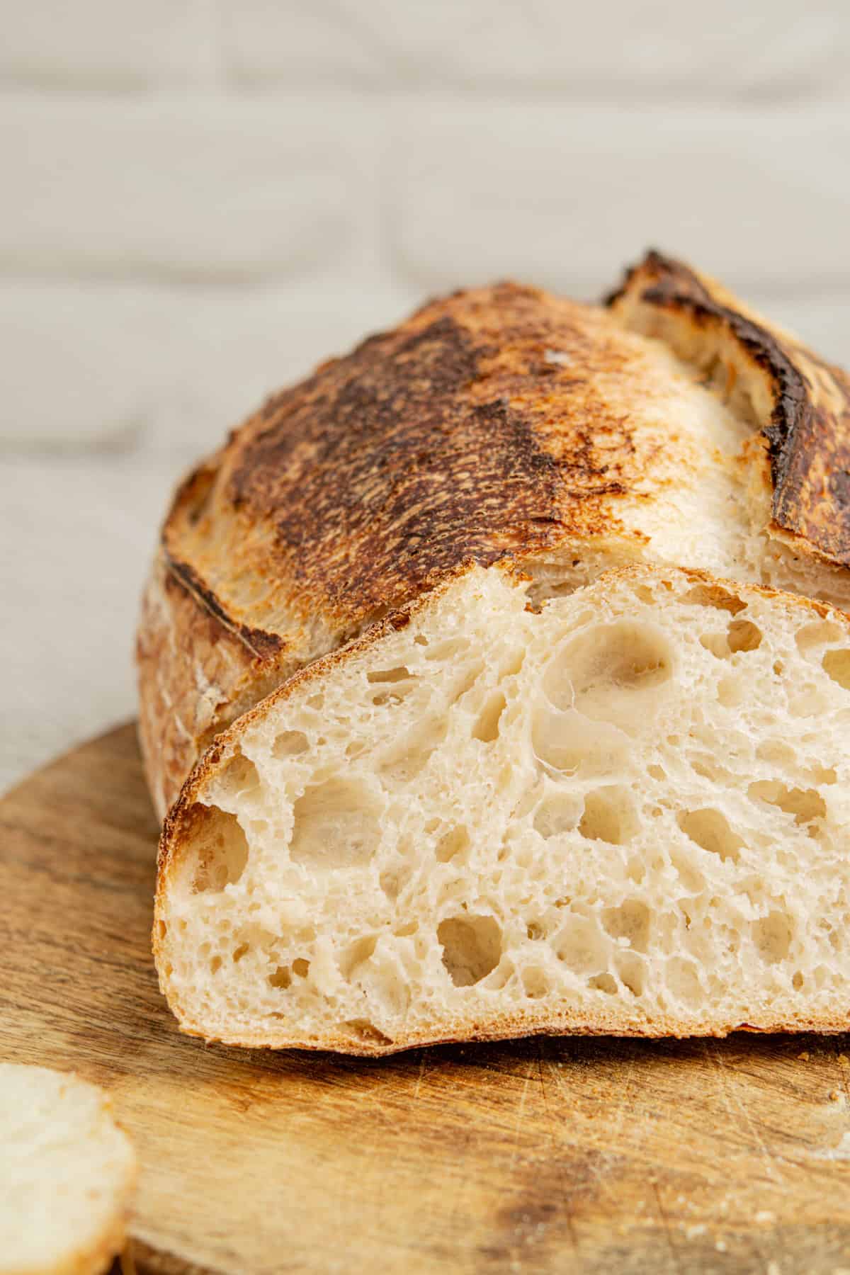 Close-up of a sliced sourdough bread on a wooden board. The bread has a golden, crispy crust and a soft, airy interior with large, irregular holes. The background is softly blurred, highlighting the texture of the bread.