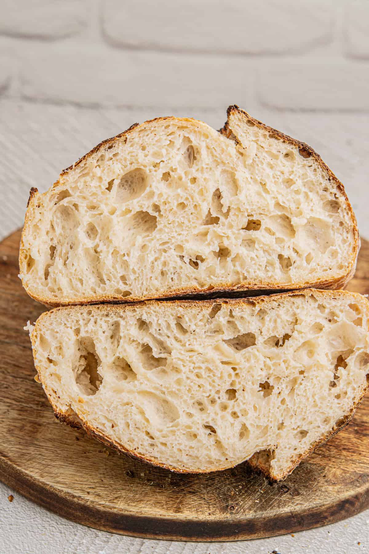 Two slices of crusty sourdough bread with a light, airy crumb are placed on a wooden cutting board. The breads golden crust and irregular holes indicate a well-baked texture. A white brick background is visible.