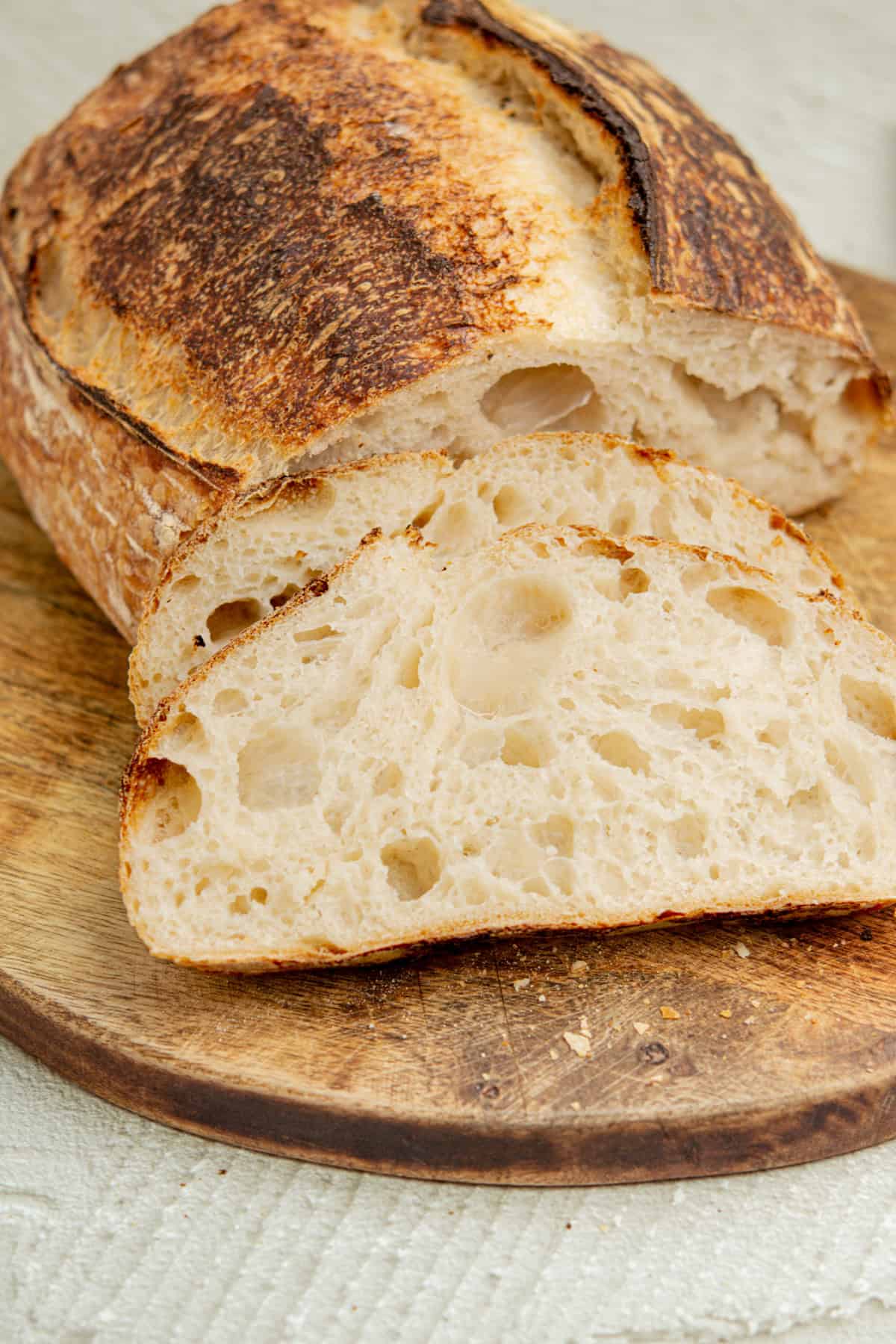 Sliced sourdough bread on a wooden board. The crust is golden brown and slightly cracked, while the interior is soft and airy with visible holes, highlighting its texture. The background is a light, neutral surface.