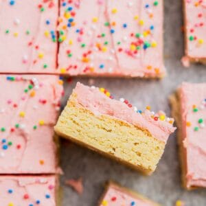 Several frosted sourdough sugar cookie bars are displayed on a surface. Each bar has a layer of pink frosting topped with colorful sprinkles. One bar is propped up to show its layers.