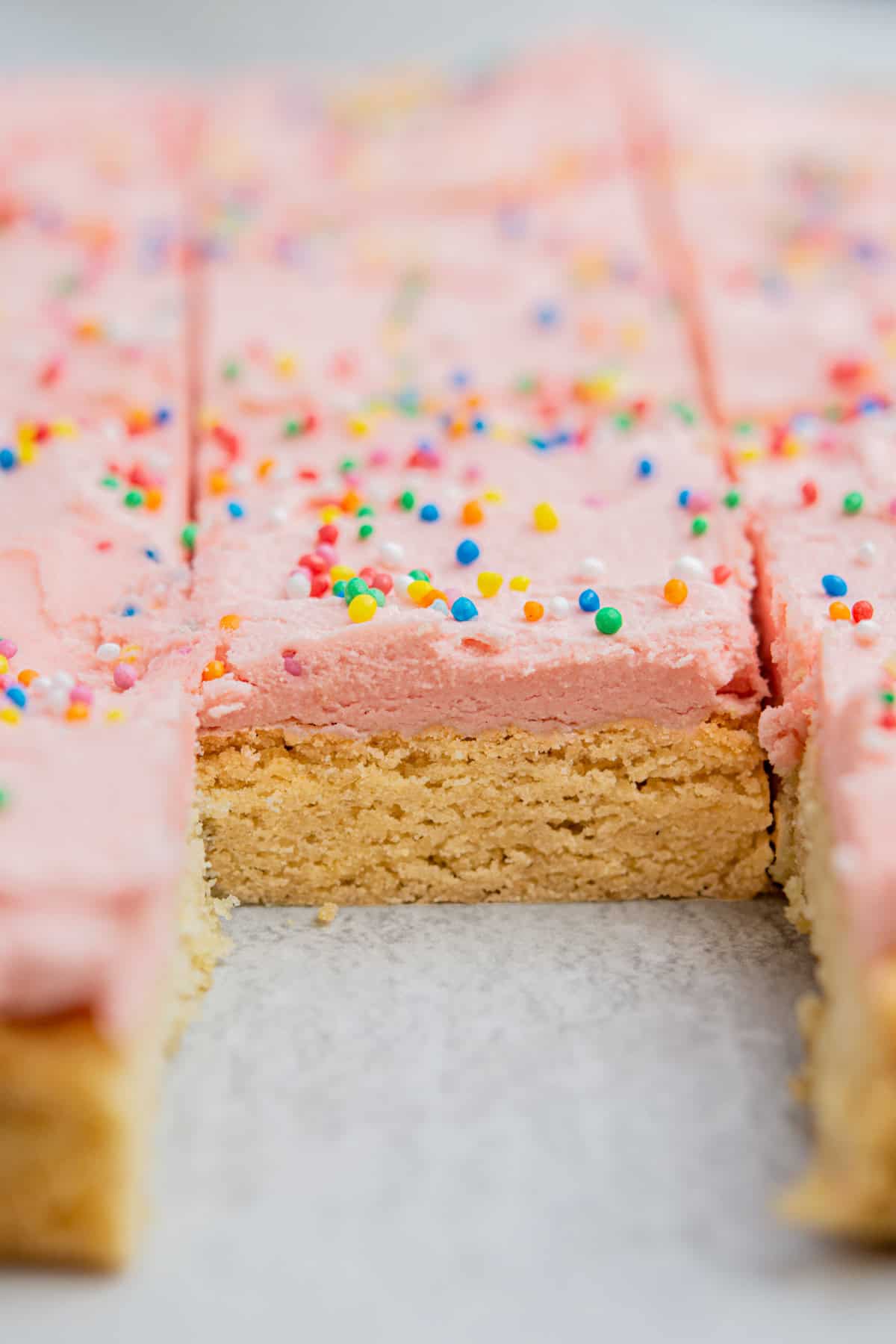 Close-up of a tray with frosted sugar cookie bars. The bars have a pink frosting top, decorated with colorful sprinkles. One bar is partially removed, showing a dense, golden-brown base. The focus is on the texture of the frosting and cookie.