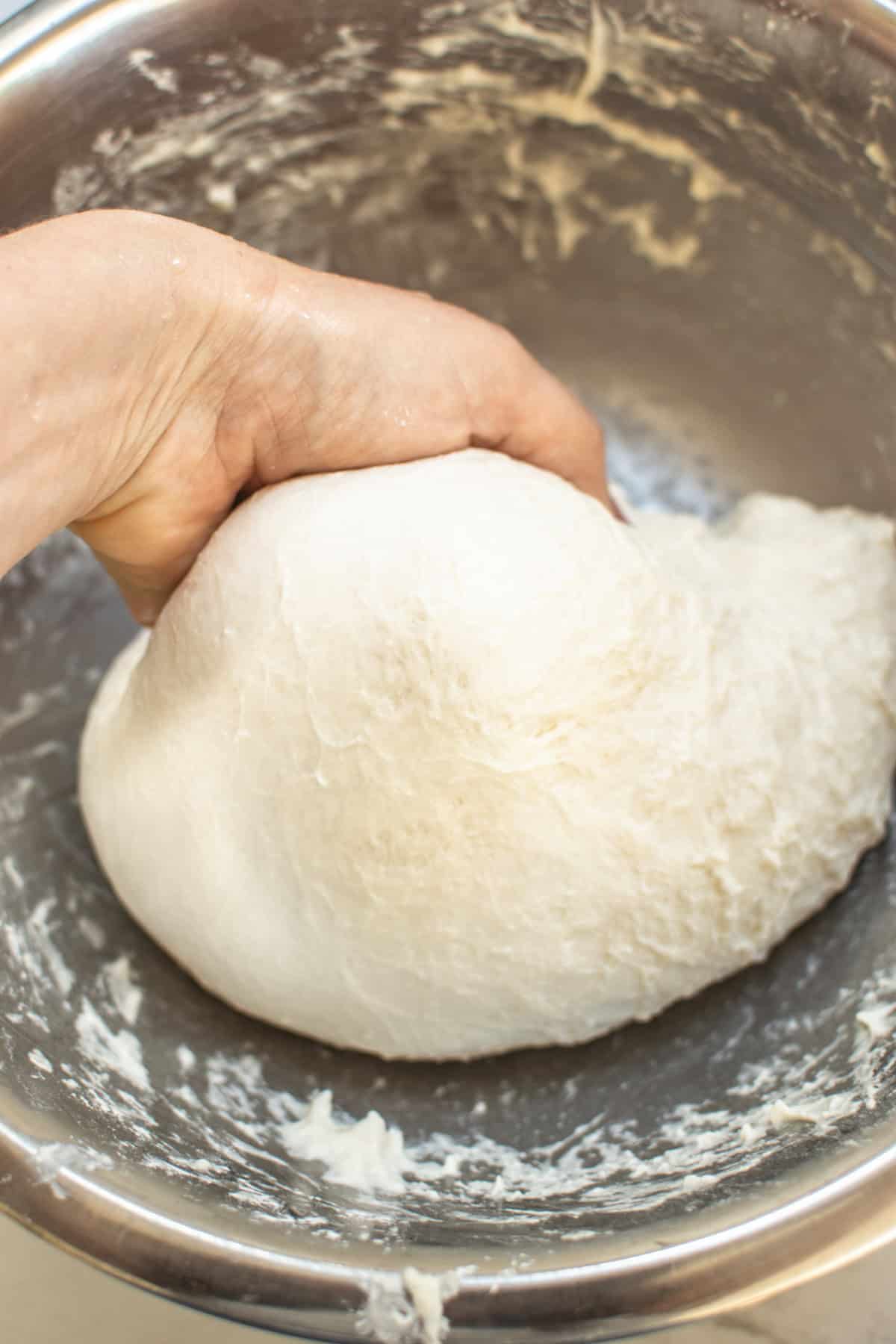 A hand kneading a soft, white dough in a metal bowl. The dough appears smooth and elastic, with some flour visible on the hand and bowl. The texture suggests its being prepared for baking or cooking.
