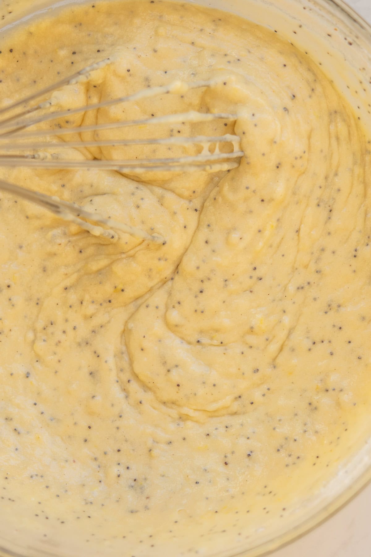 A whisk is mixing a thick, yellow batter with small black specks in a bowl. The batter appears creamy and smooth, likely a cake or muffin mixture with added ingredients like poppy seeds.