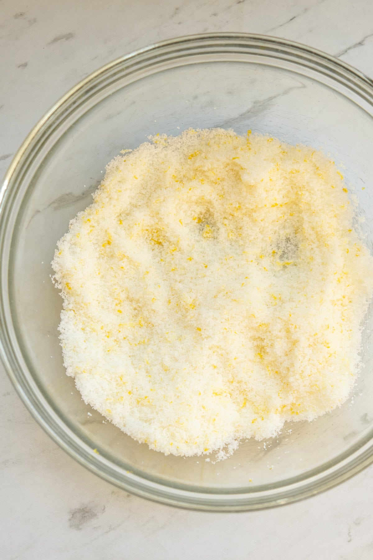 A glass bowl filled with a mixture of granulated sugar and lemon zest sits on a light marble surface. The sugar appears fluffy with hints of yellow zest distributed throughout.