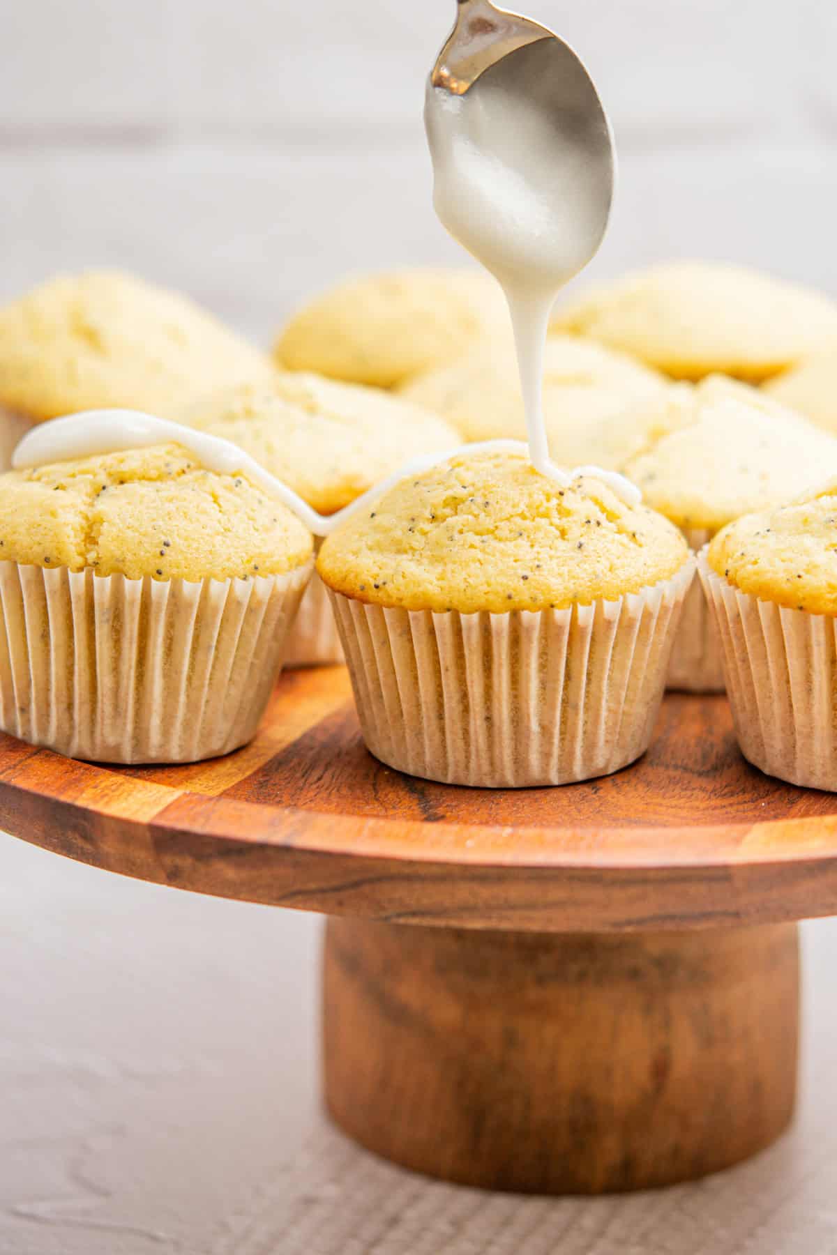 A wooden stand displays freshly baked muffins in paper wrappers. A spoon drizzles white icing over one muffin, adding a sweet touch. The background is softly blurred, highlighting the muffins.