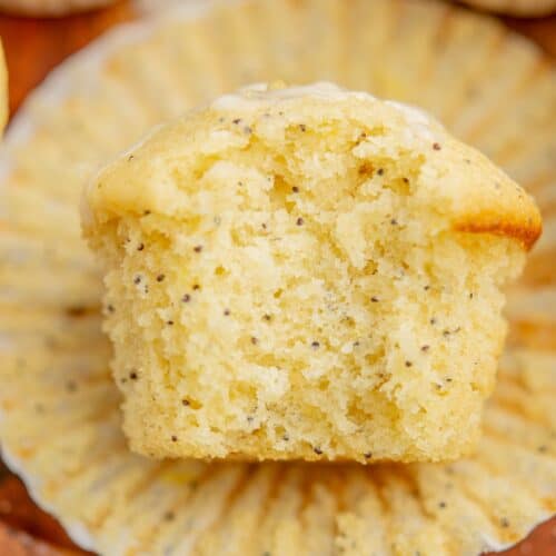 Close-up of a moist sourdough lemon poppy seed muffin, cut in half, revealing its fluffy texture and speckled with poppy seeds. The muffin rests on an unfolded paper liner, showcasing its golden, slightly crumbly top.