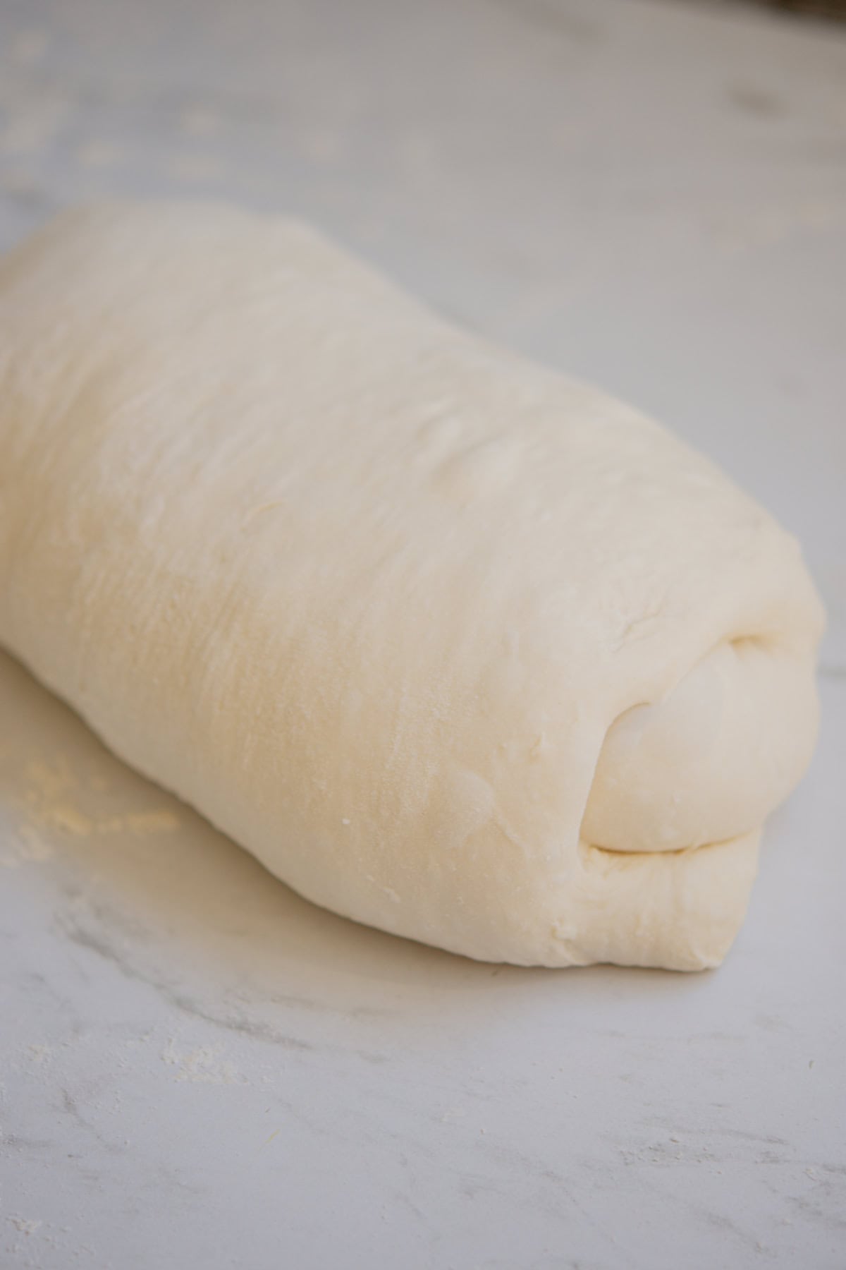 A close-up of a neatly rolled piece of dough resting on a lightly floured marble surface, ready for baking. The dough appears soft and smooth, with its edges tucked in carefully.