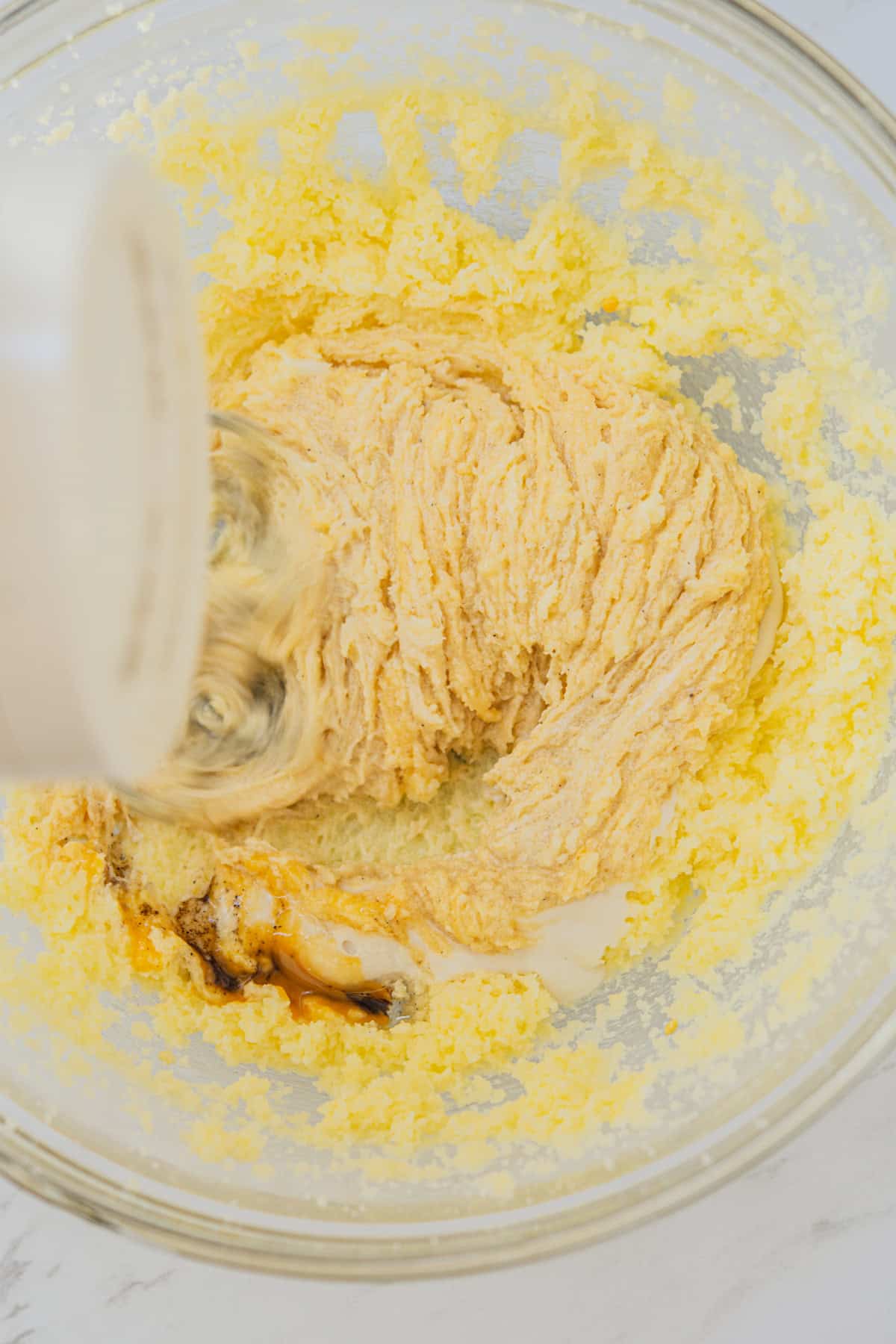 A mixing bowl containing a creamy, yellow batter being blended with a hand mixer. Vanilla extract is visible in the batter, giving it a swirled appearance. The background is a light, marble surface.