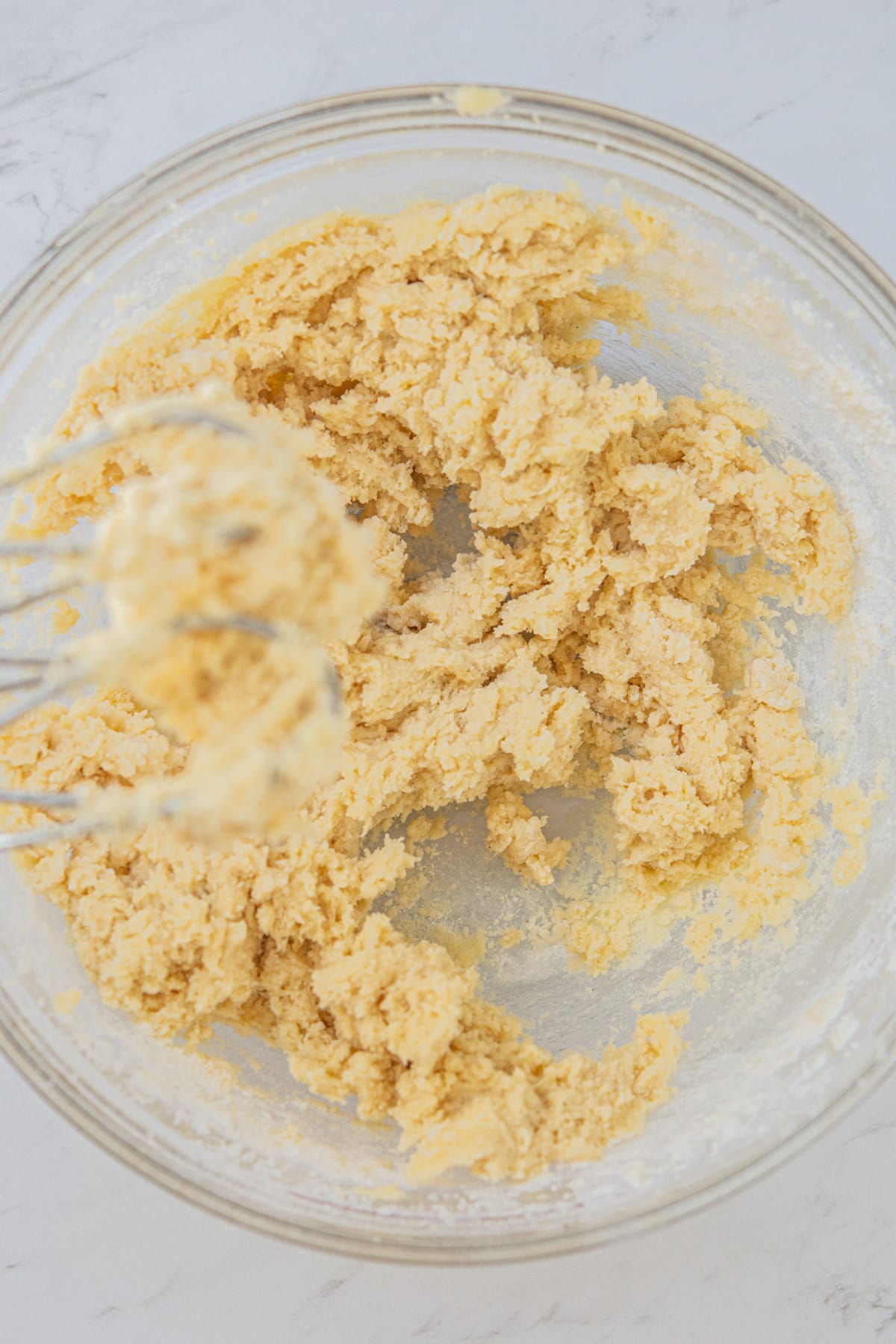 A glass bowl containing a thick, creamy mixture of cookie dough. A whisk is partially visible, coated with the dough. The bowl is placed on a white countertop.