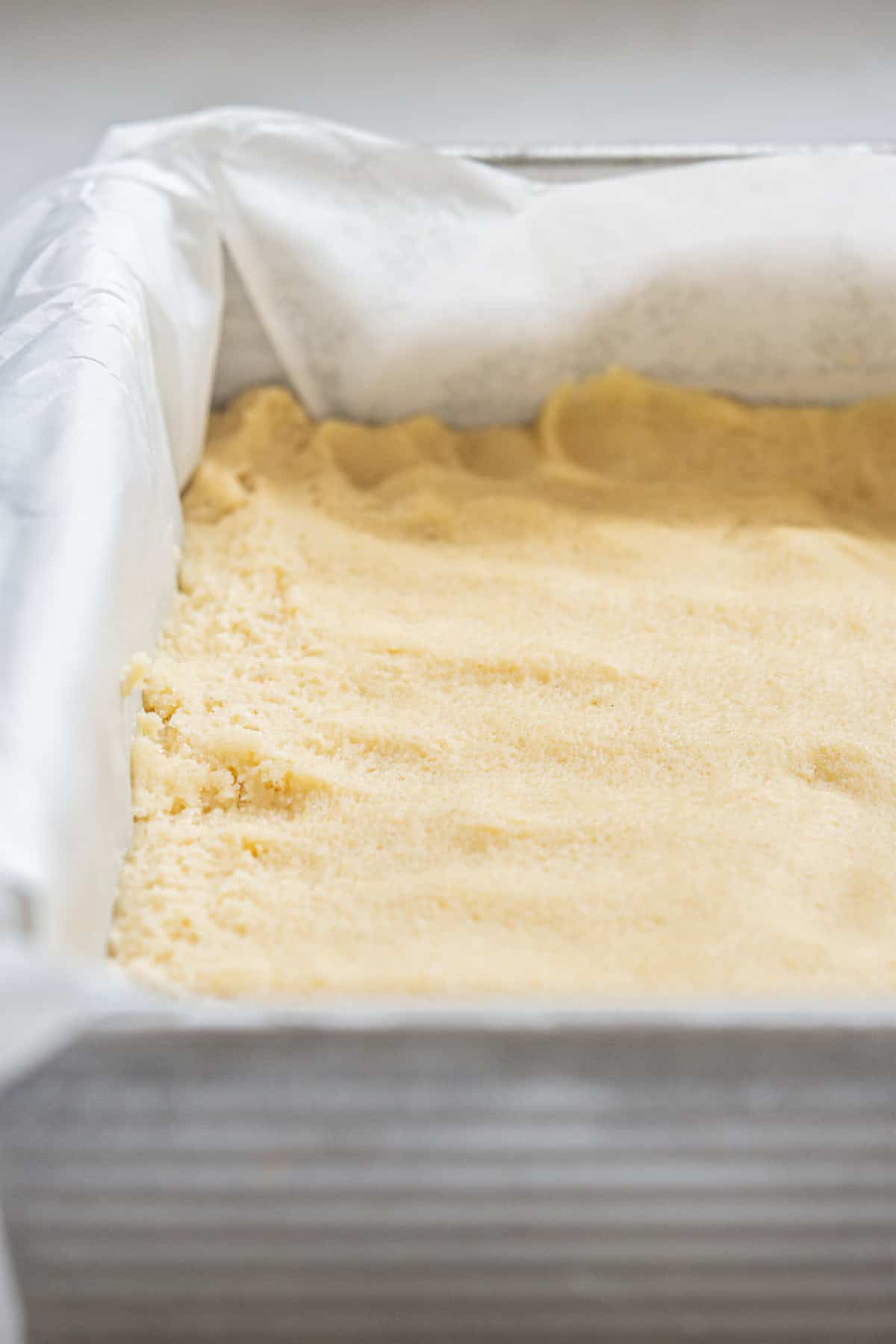 A rectangular baking pan lined with parchment paper holds a layer of unbaked shortbread dough, evenly spread and ready for the oven. The dough appears smooth and creamy in texture.