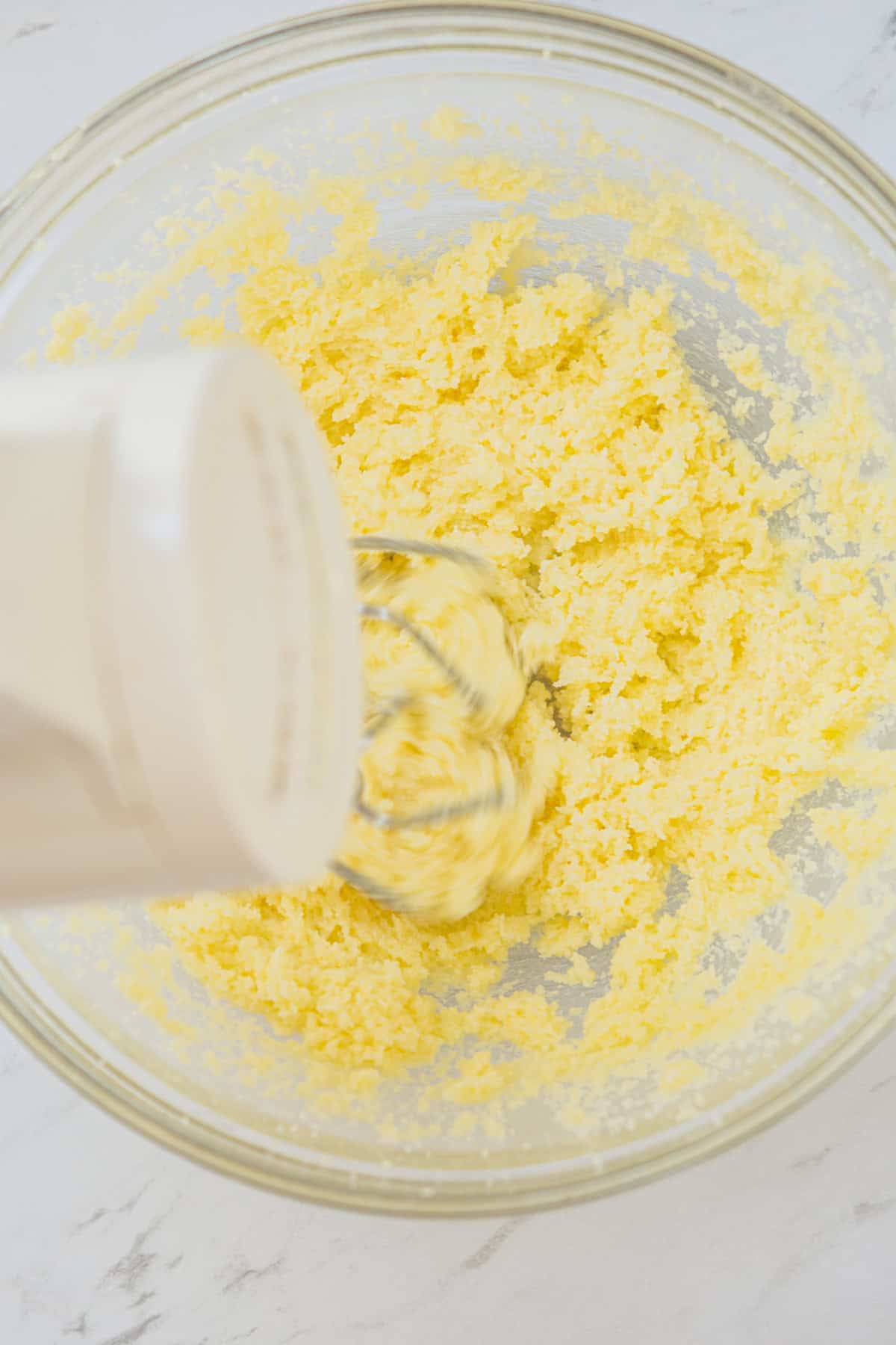 A close-up of a hand mixer blending butter and sugar in a transparent glass bowl. The mixture is light and fluffy, showing a creamy texture. The bowl rests on a marble countertop.