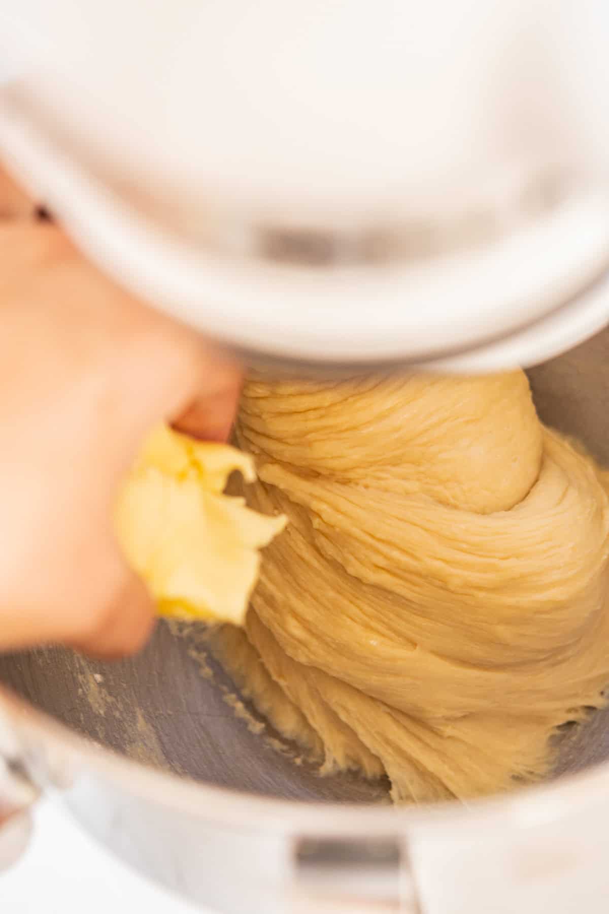 A hand adds butter to dough while it mixes in a stand mixer. The dough appears smooth and elastic, indicating it is being kneaded. The action suggests the process of making bread or pastries.