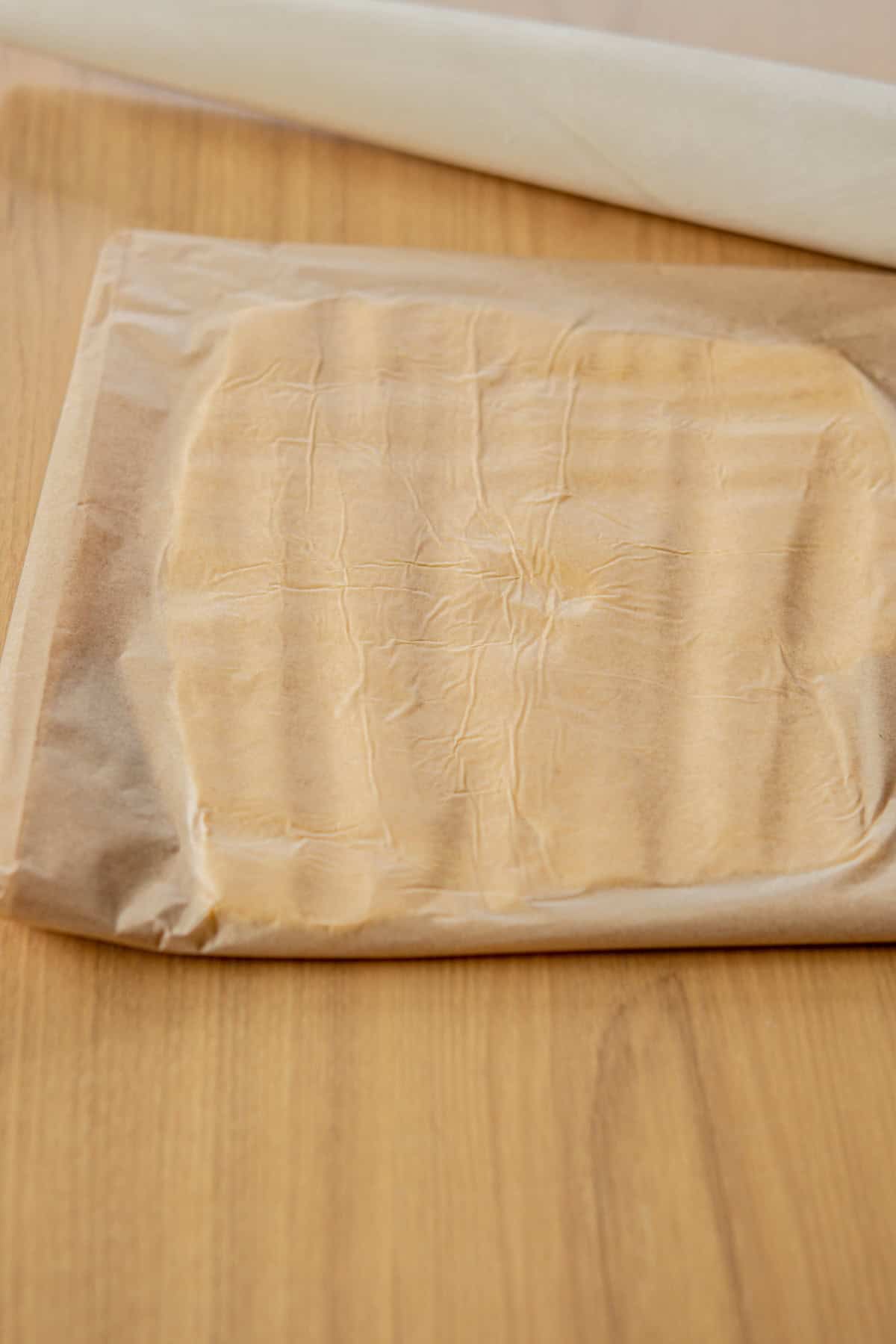 Brown parchment paper with dough inside lies on a wooden surface. A white rolling pin is partially visible at the top of the image.