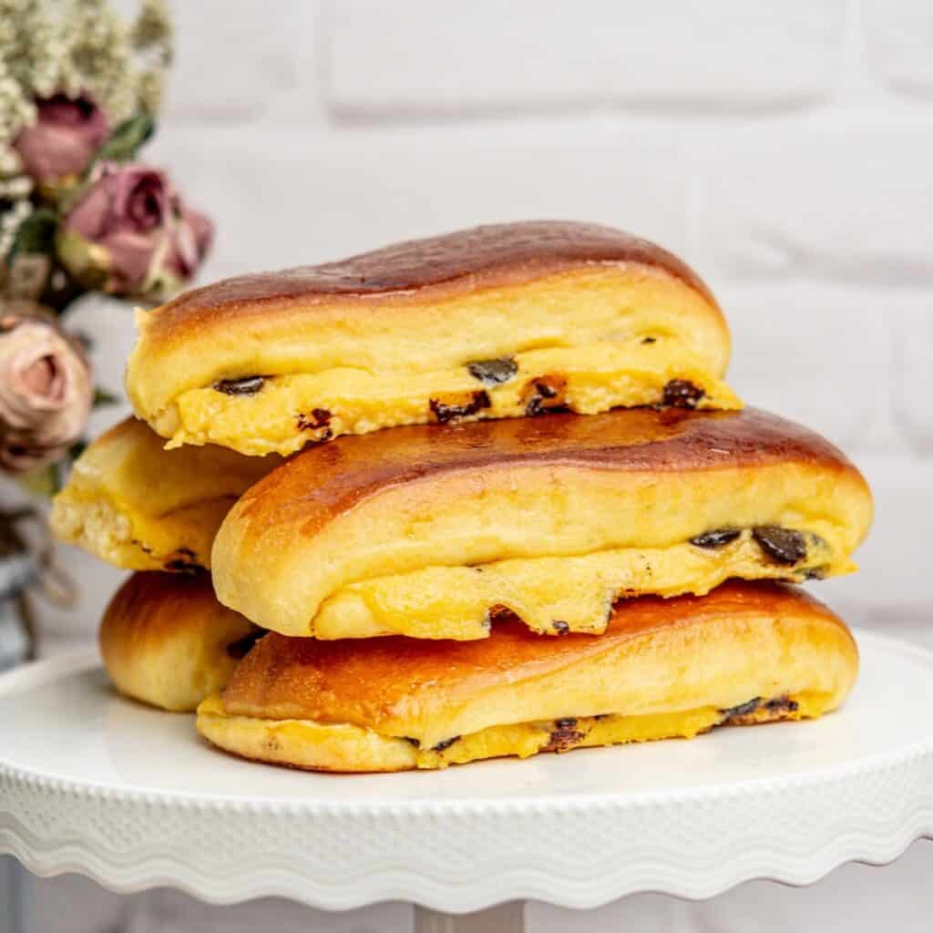 Three sweet brioche suisse filled with custard and raisins are stacked on a white cake stand. The pastries have a golden-brown crust and are placed against a white brick wall background, with dried flowers visible in the corner.