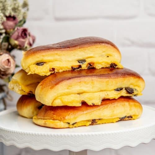 Three sweet brioche suisse filled with custard and raisins are stacked on a white cake stand. The pastries have a golden-brown crust and are placed against a white brick wall background, with dried flowers visible in the corner.