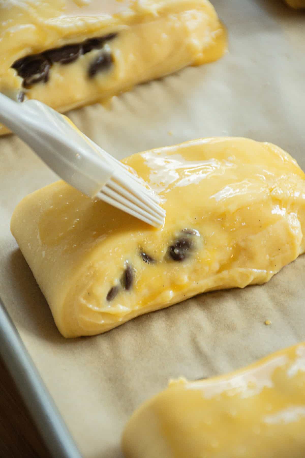Close-up of a pastry brush applying egg wash to unbaked chocolate pastries on a parchment-lined baking sheet. The dough is shiny, light yellow, and glossy, with visible chocolate pieces inside.