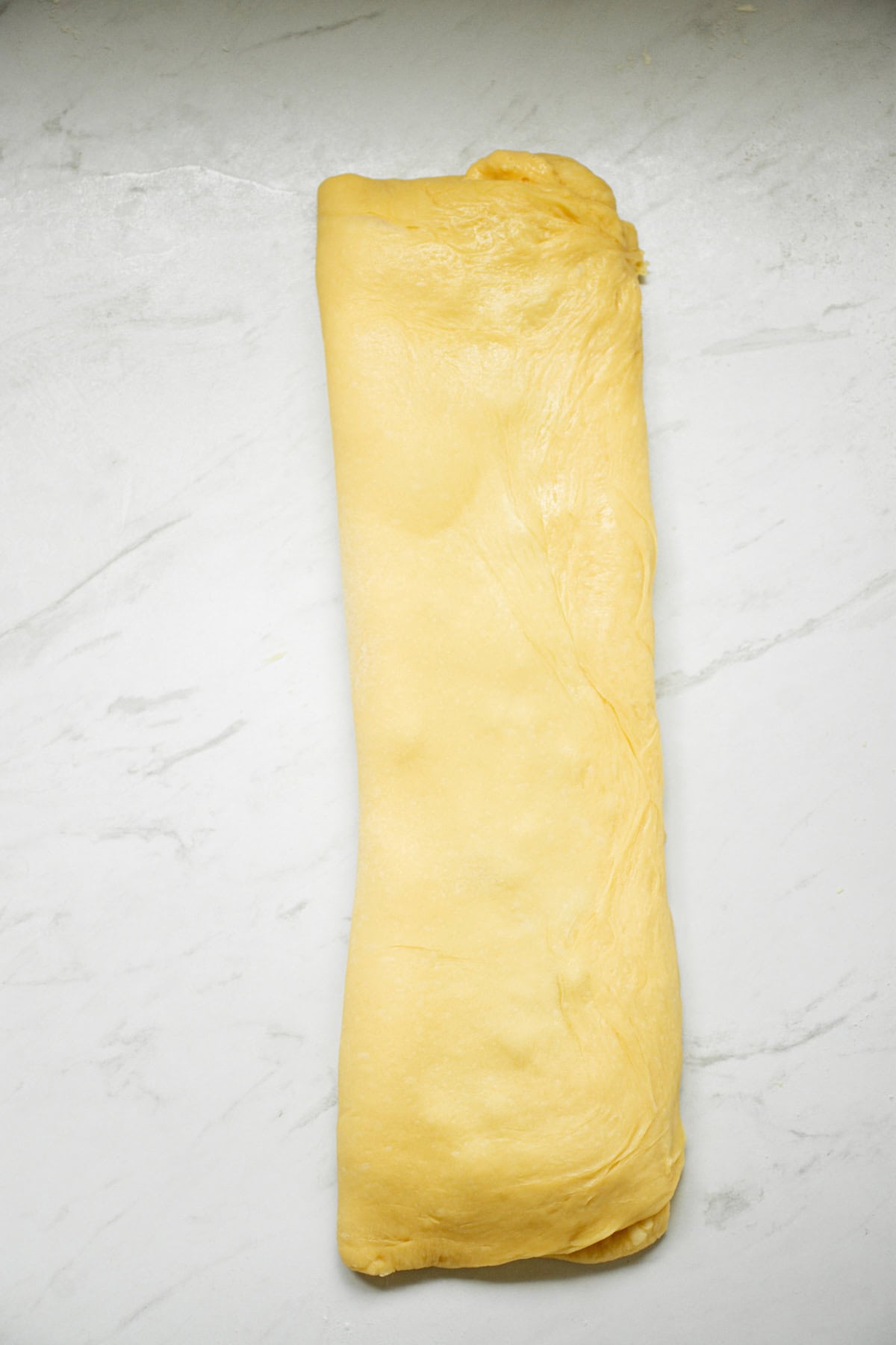 A neatly rolled piece of yellow dough on a white marble surface. The dough appears smooth and evenly shaped, suggesting it is ready for further preparation in cooking or baking.