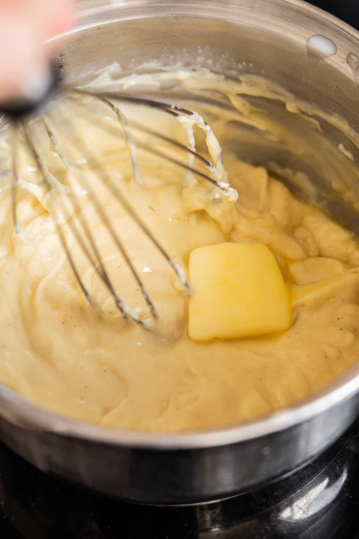 A whisk stirs creamy mashed potatoes with a melting pat of butter in a stainless steel pot.