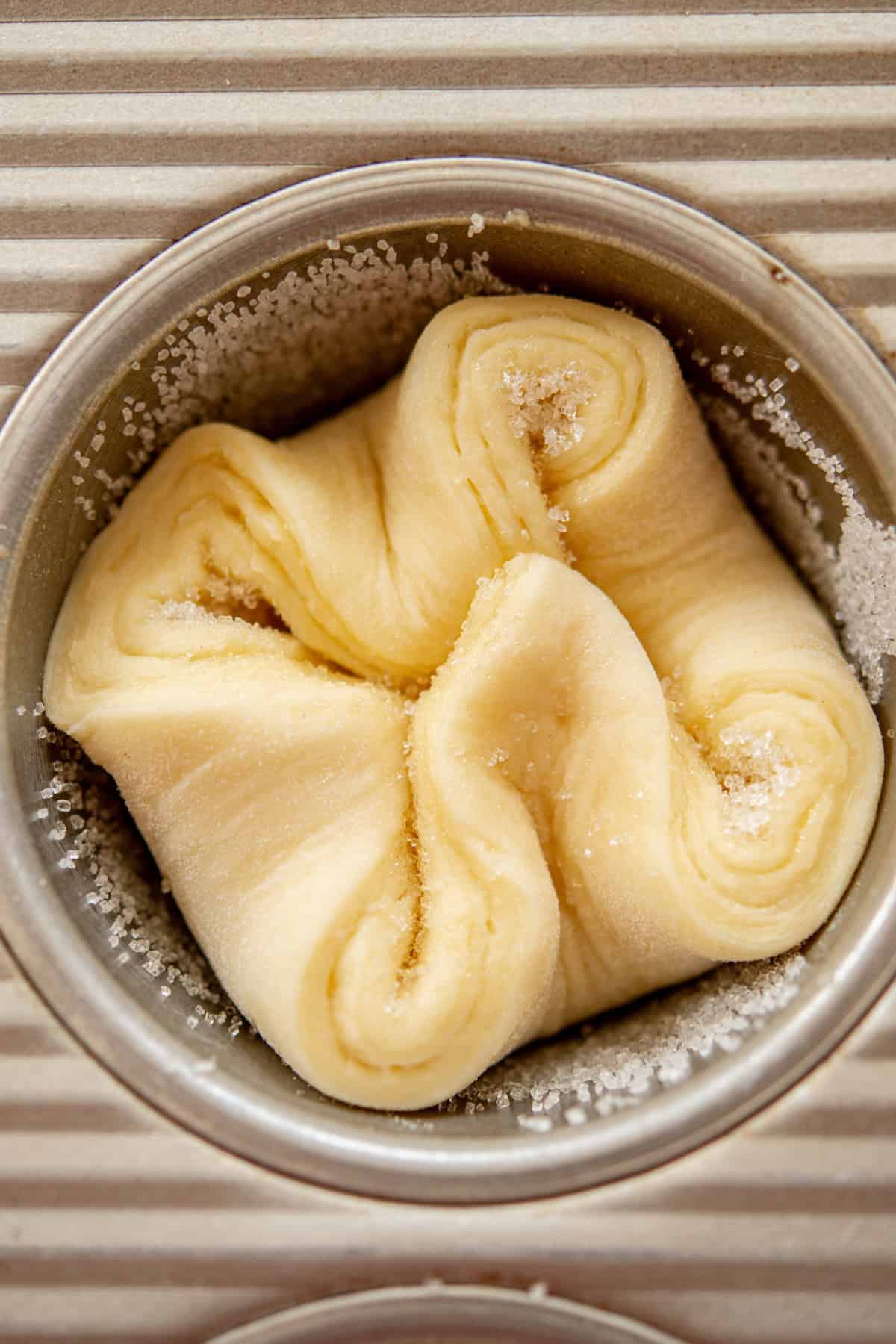 A piece of folded pastry dough with sugar sprinkled on top, placed in a small metal dish. The dough has been arranged in a decorative, layered shape. The background features a textured, ribbed surface.