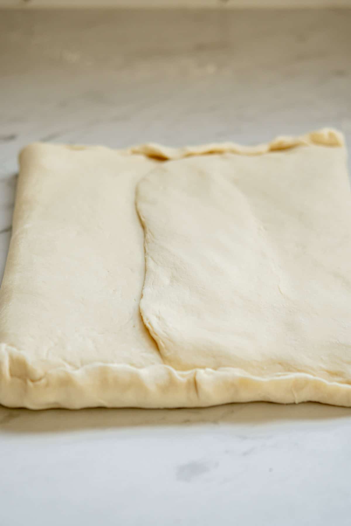 A close-up of a neatly folded sheet of raw puff pastry dough on a light marble countertop. The dough is golden and smooth, with slight creases where it has been folded.