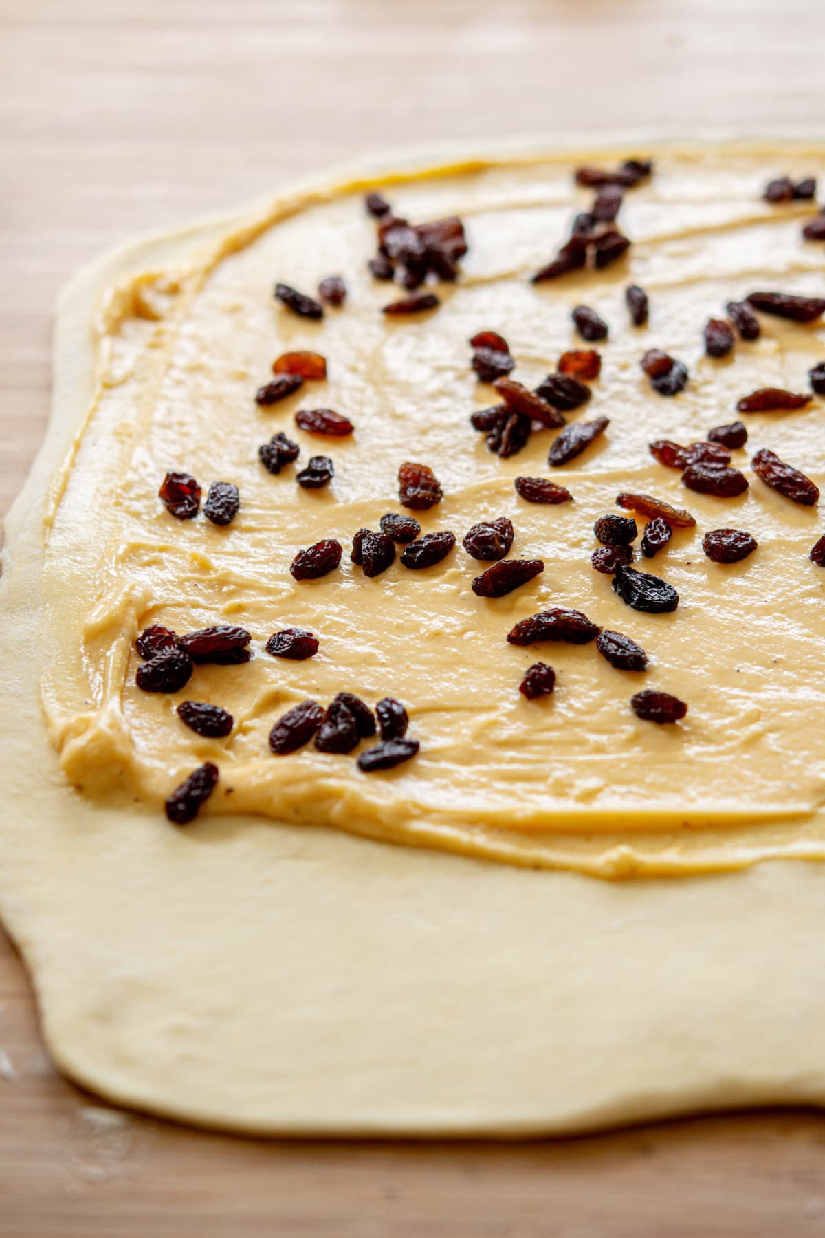 Rolled-out dough spread with a creamy yellow mixture and sprinkled with raisins, placed on a wooden surface.