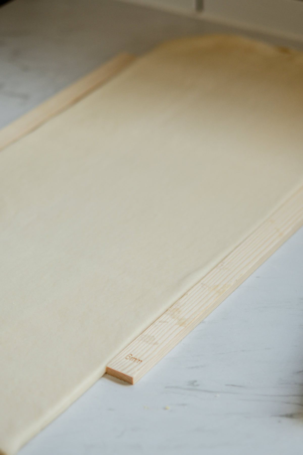 A sheet of rolled pastry dough placed on a light wooden board, set on a marble surface. The dough is smooth and evenly spread, with a rolling pin partially visible at the edge.
