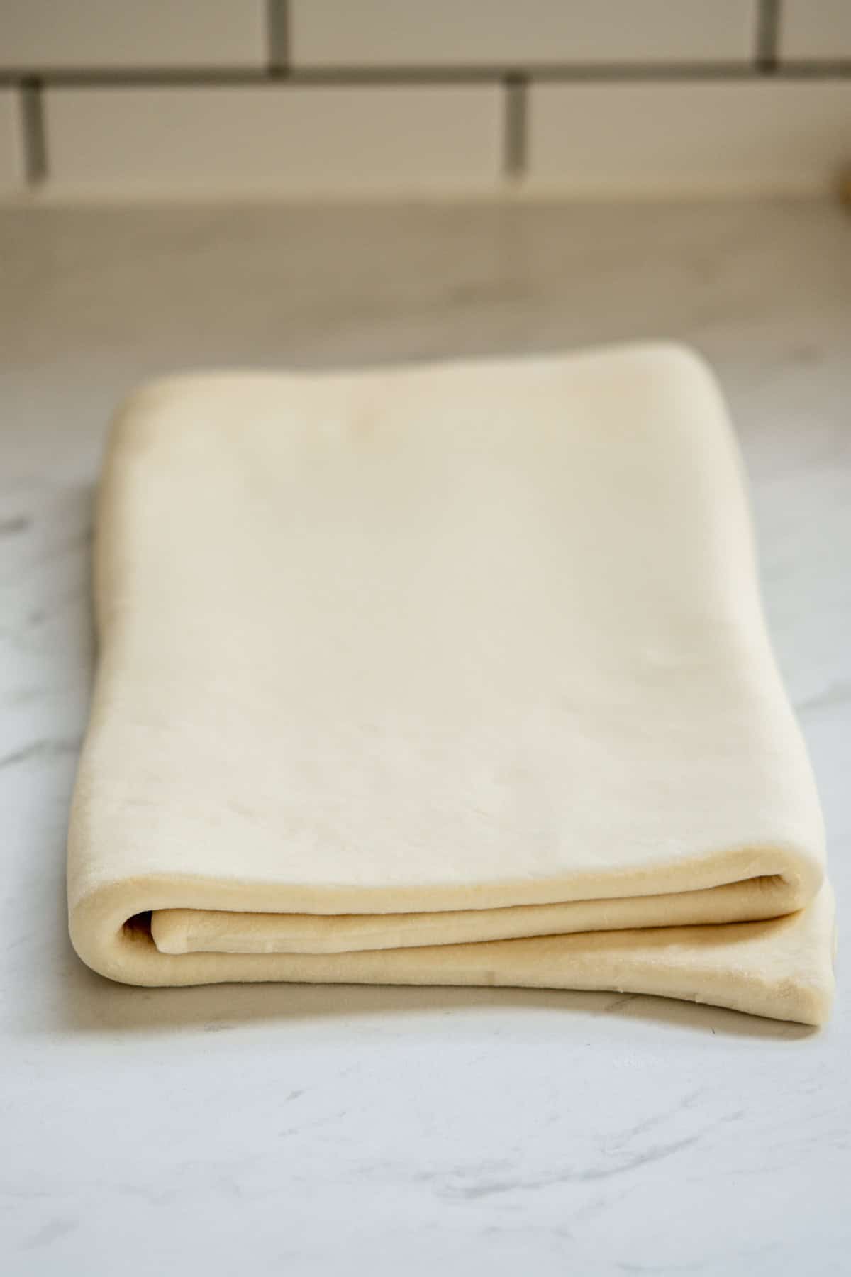 A neatly folded sheet of light cream-colored puff pastry rests on a marble countertop, with white subway tiles in the background.