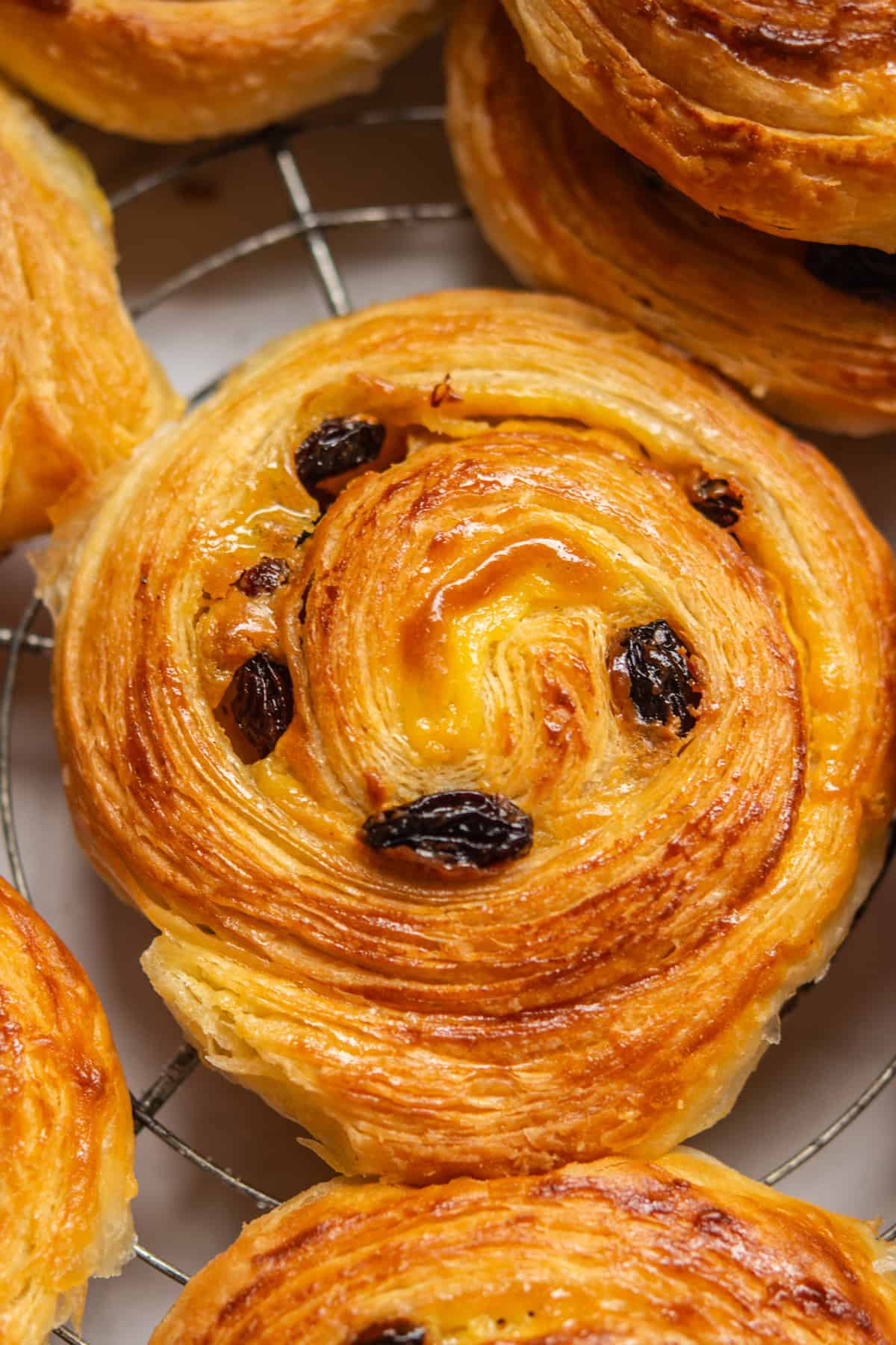 Close-up of a golden-brown, flaky pastry swirl with raisins and a glossy finish, resting on a wire rack. Other pastries are partially visible in the background.