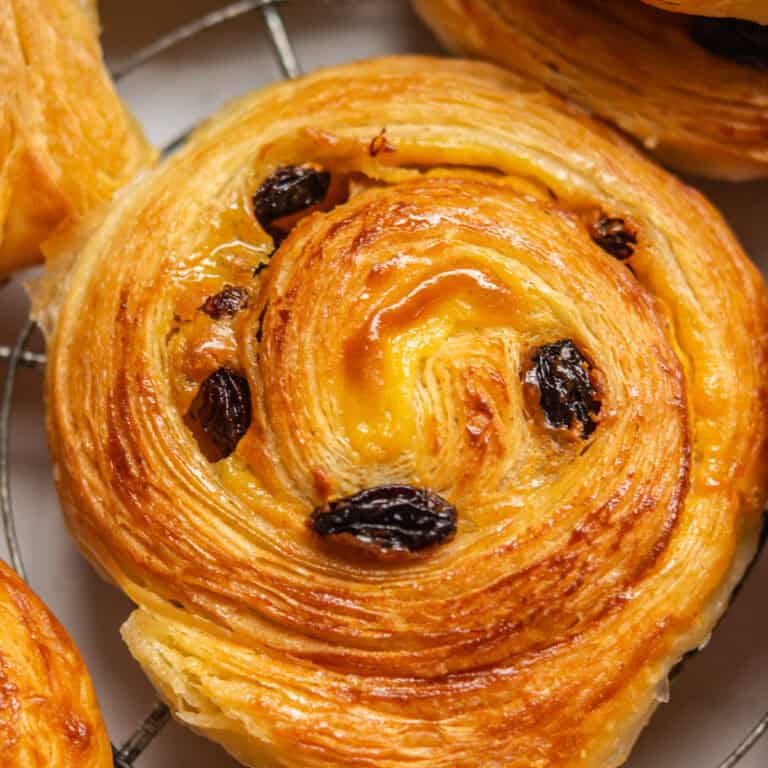 Close-up of a freshly baked raisin swirl pastry on a cooling rack. The golden-brown layers are flaky and glossy, with raisins embedded in the swirl pattern.
