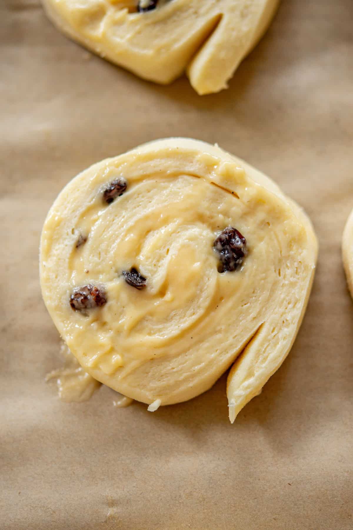 Close-up of an uncooked cinnamon roll on parchment paper. The roll is swirled with visible raisins embedded in the dough. The texture appears soft and doughy, ready for baking.